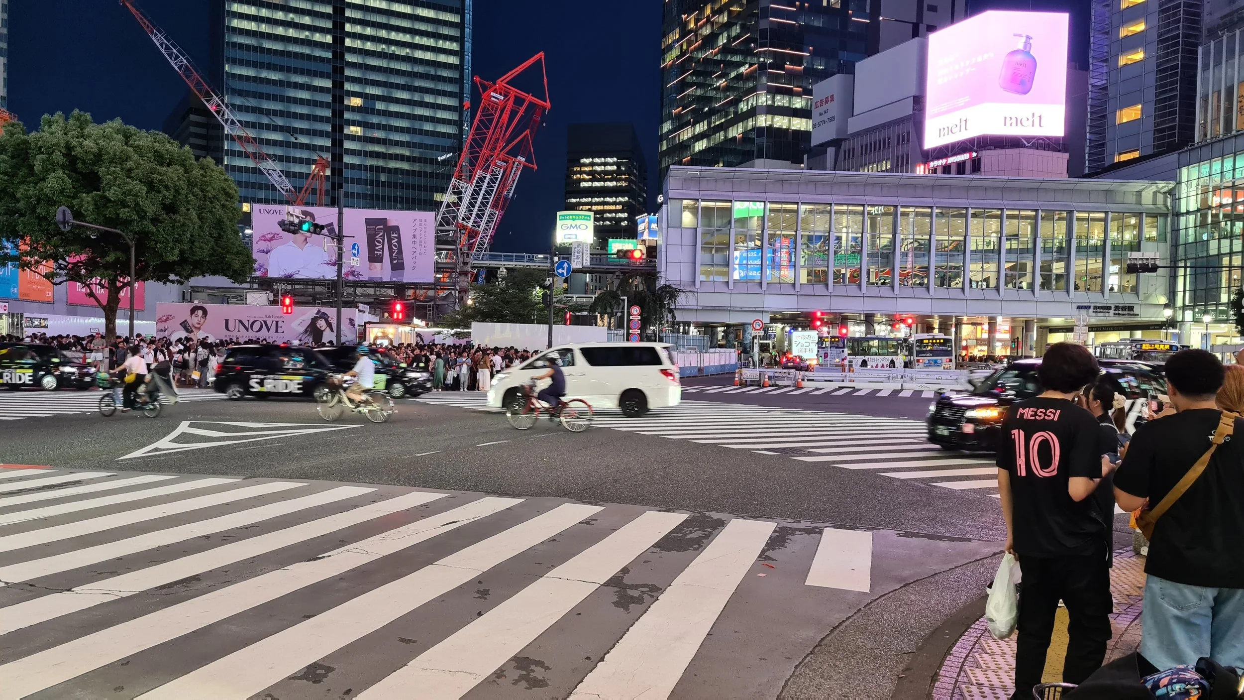 A busy city intersection at night with pedestrians waiting at the crosswalk, vehicles, and bicycles crossing. Tall buildings with illuminated billboards and advertisements surround the scene.