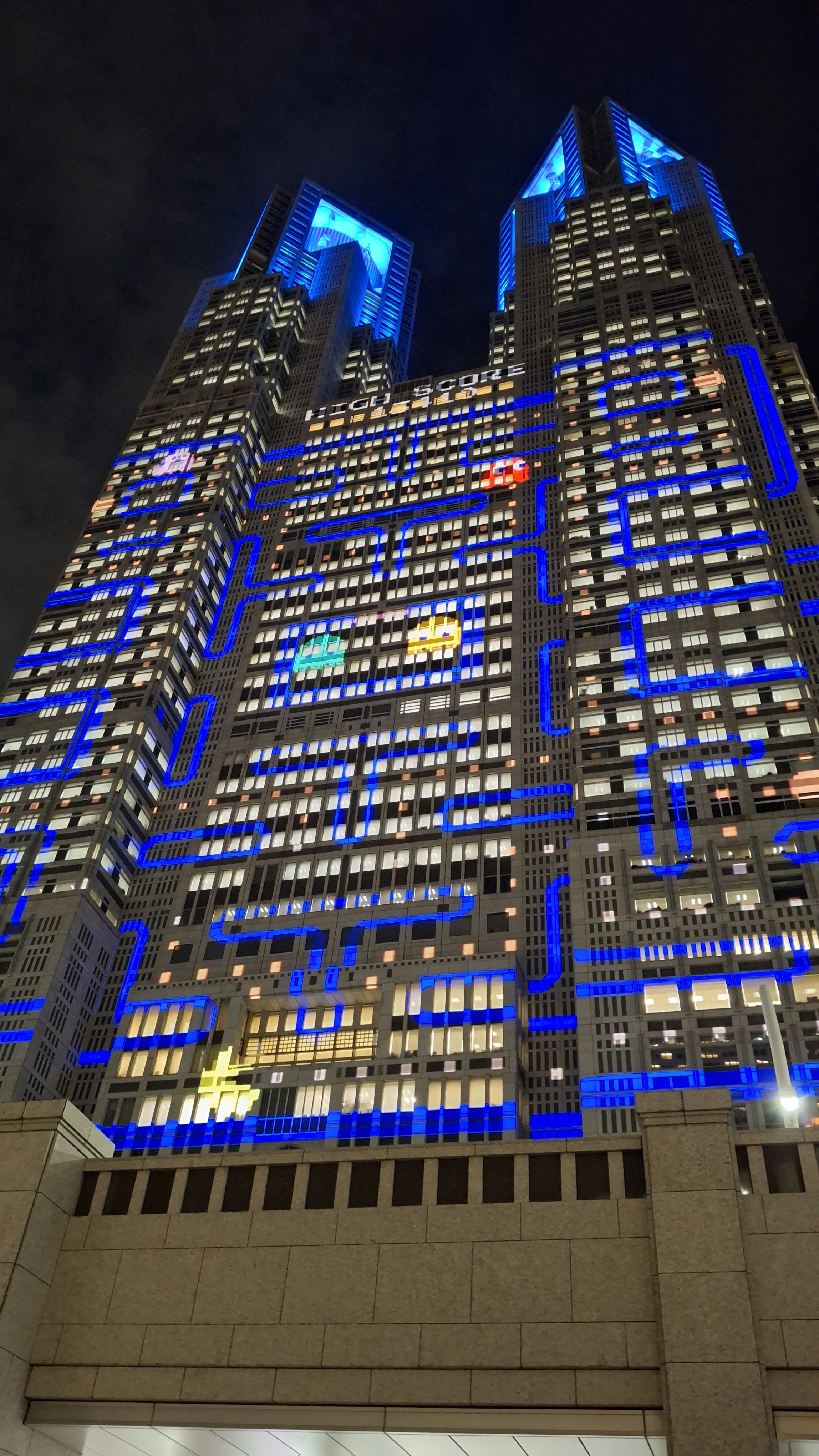 Night view of a tall building decorated with bright blue, yellow, and light pink lights designed to look like a video game with pixel art style. The building features a Pac-Man theme with Pac-Man and ghosts, as well as the phrase 'HIGH SCORE' display
