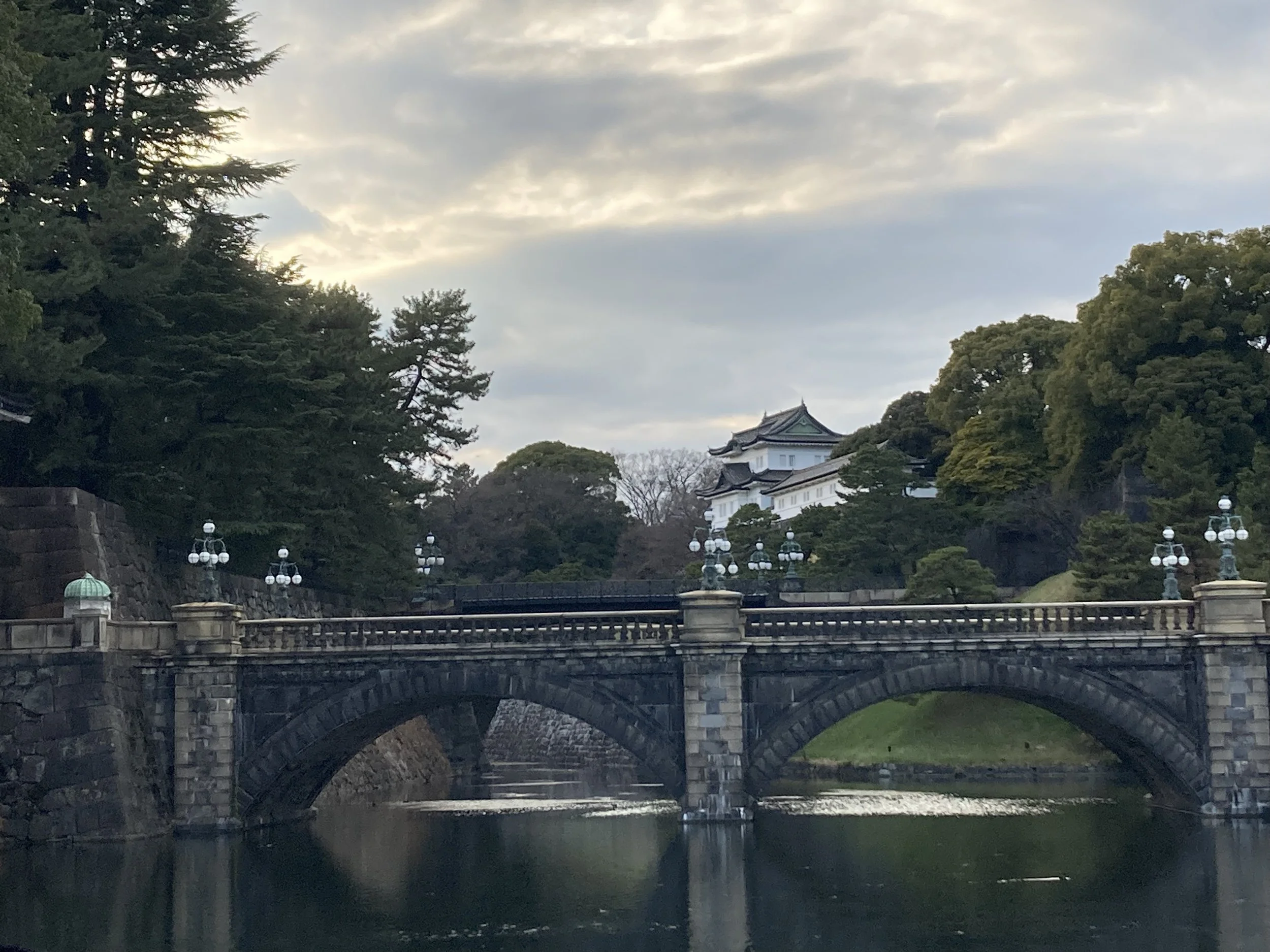 A stone bridge over a river with a white traditional Japanese building on a hill surrounded by green trees under a cloudy sky.
