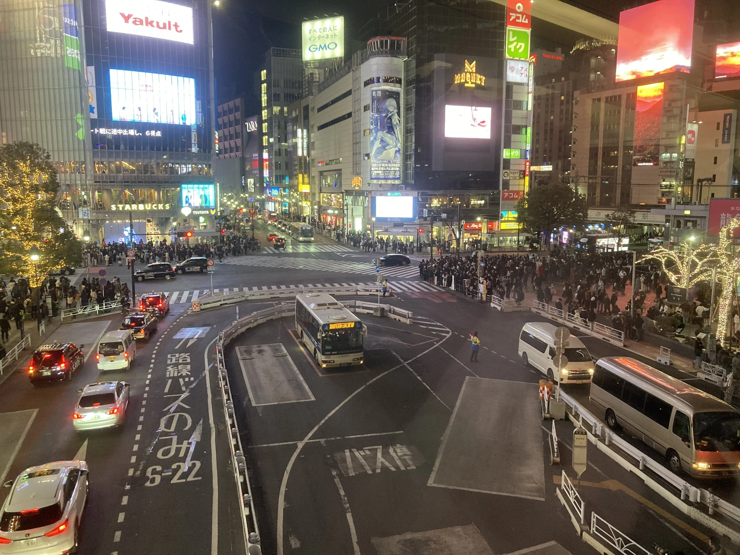 Nighttime view of a busy city intersection with multiple cars and buses, illuminated billboards, and crowds of pedestrians. Recognizable stores include Starbucks and Tsutaya.