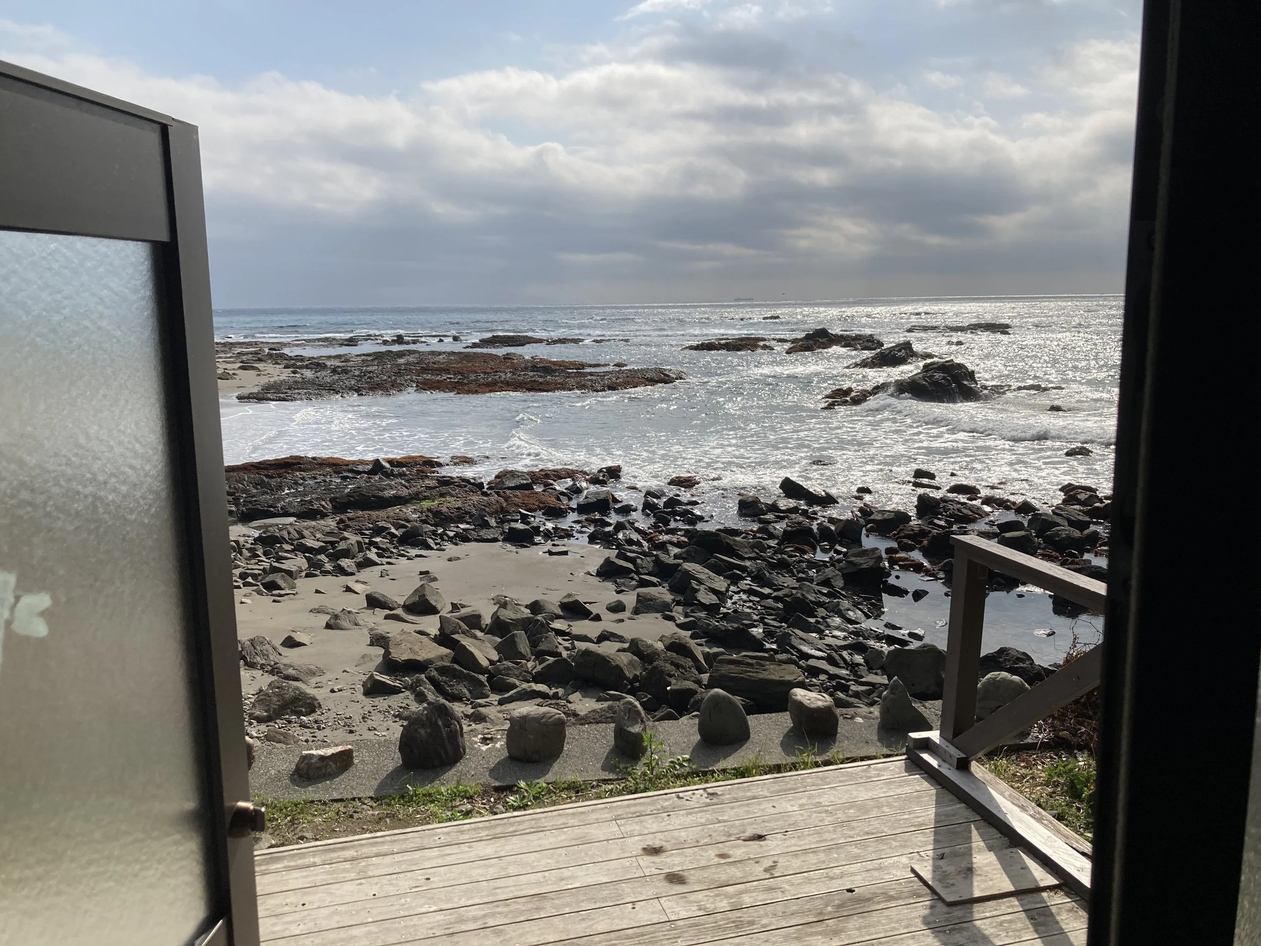 View of a rocky beach through an open door with a wooden deck, ocean waves, and a partly cloudy sky.