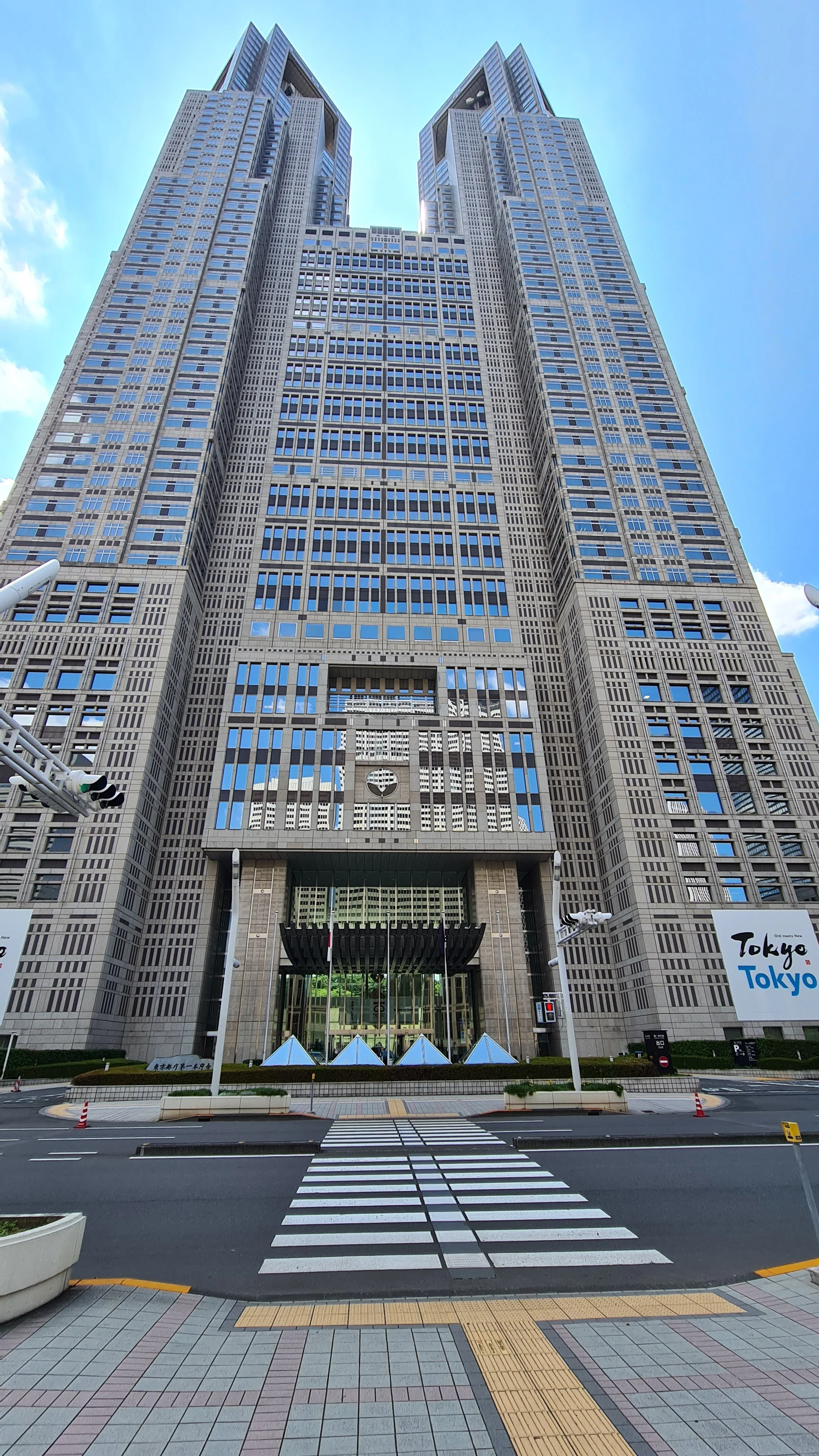A tall modern skyscraper with a unique design, featuring two tower-like structures connected in the middle, situated at a street corner with crosswalks and street lights in front.