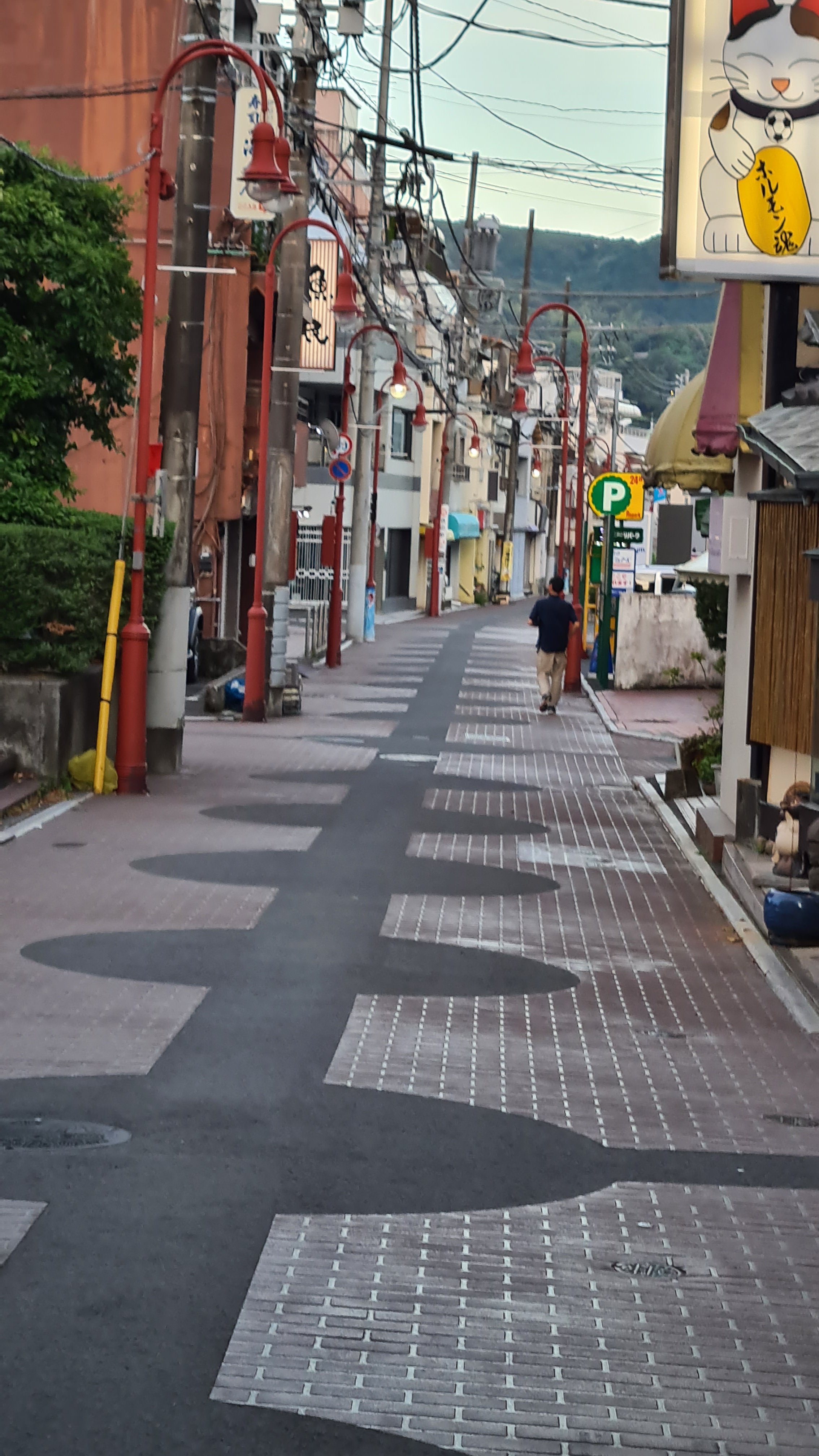Empty street with patterned pavement, red street lamps, overhead wiring, and small shops, with a person walking away in the distance.