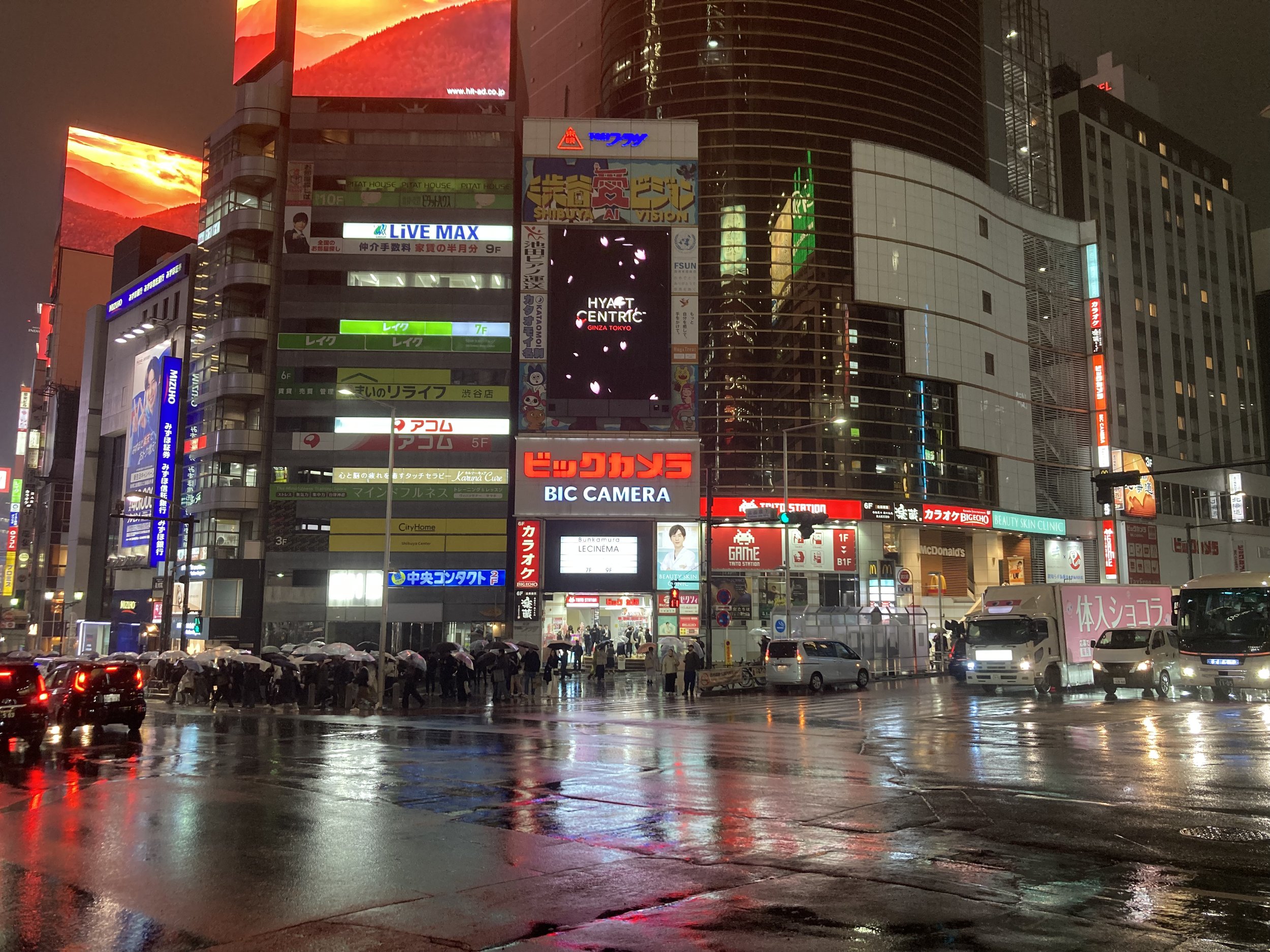 Nighttime city scene featuring wet streets reflecting colorful neon signs and advertisements on tall buildings, with a crowd of people holding umbrellas and cars driving through the area.