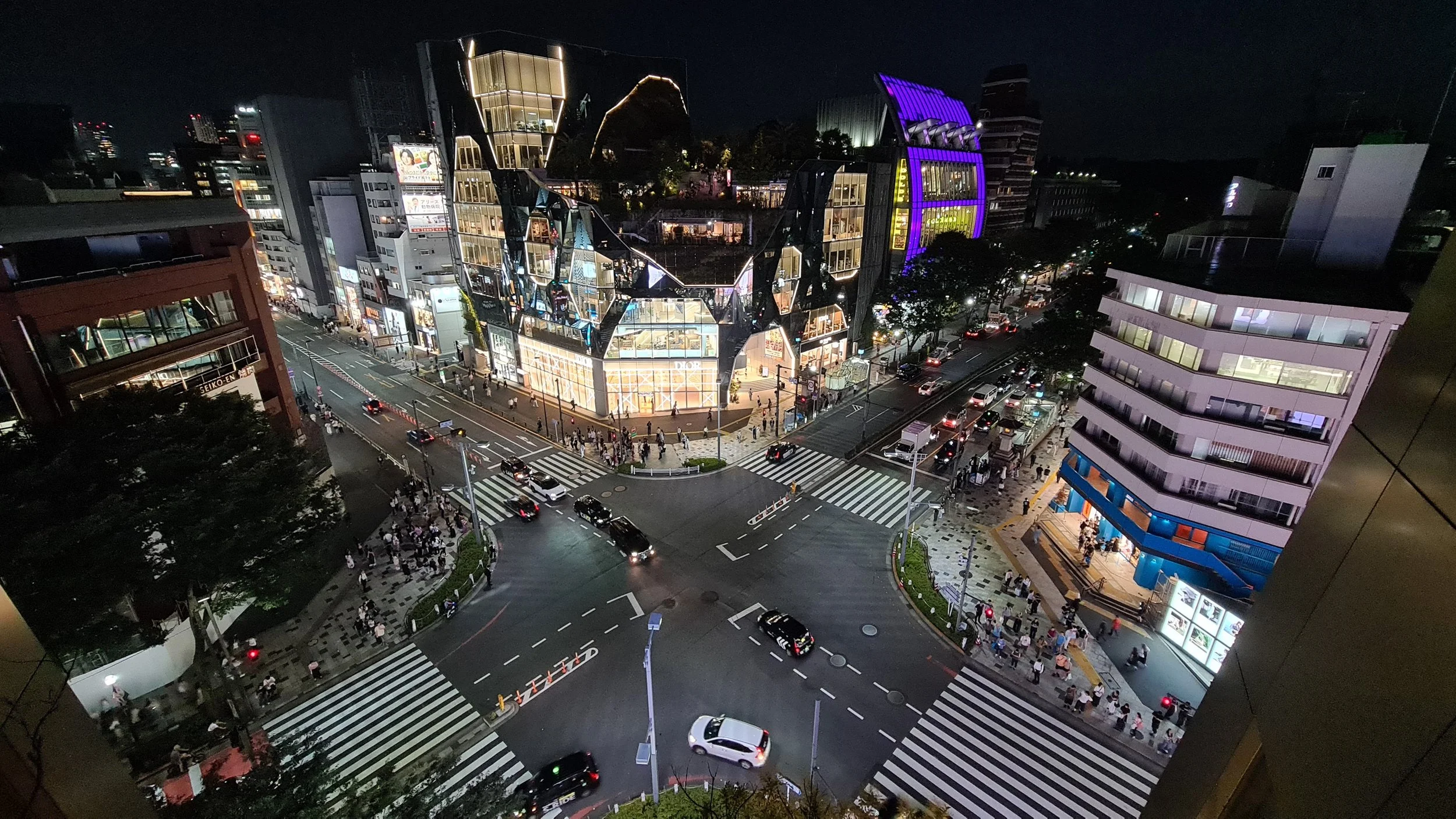Nighttime cityscape with illuminated modern buildings, busy intersections with cars and pedestrians, and a distinctive multi-angled glass building at a busy crossroad.