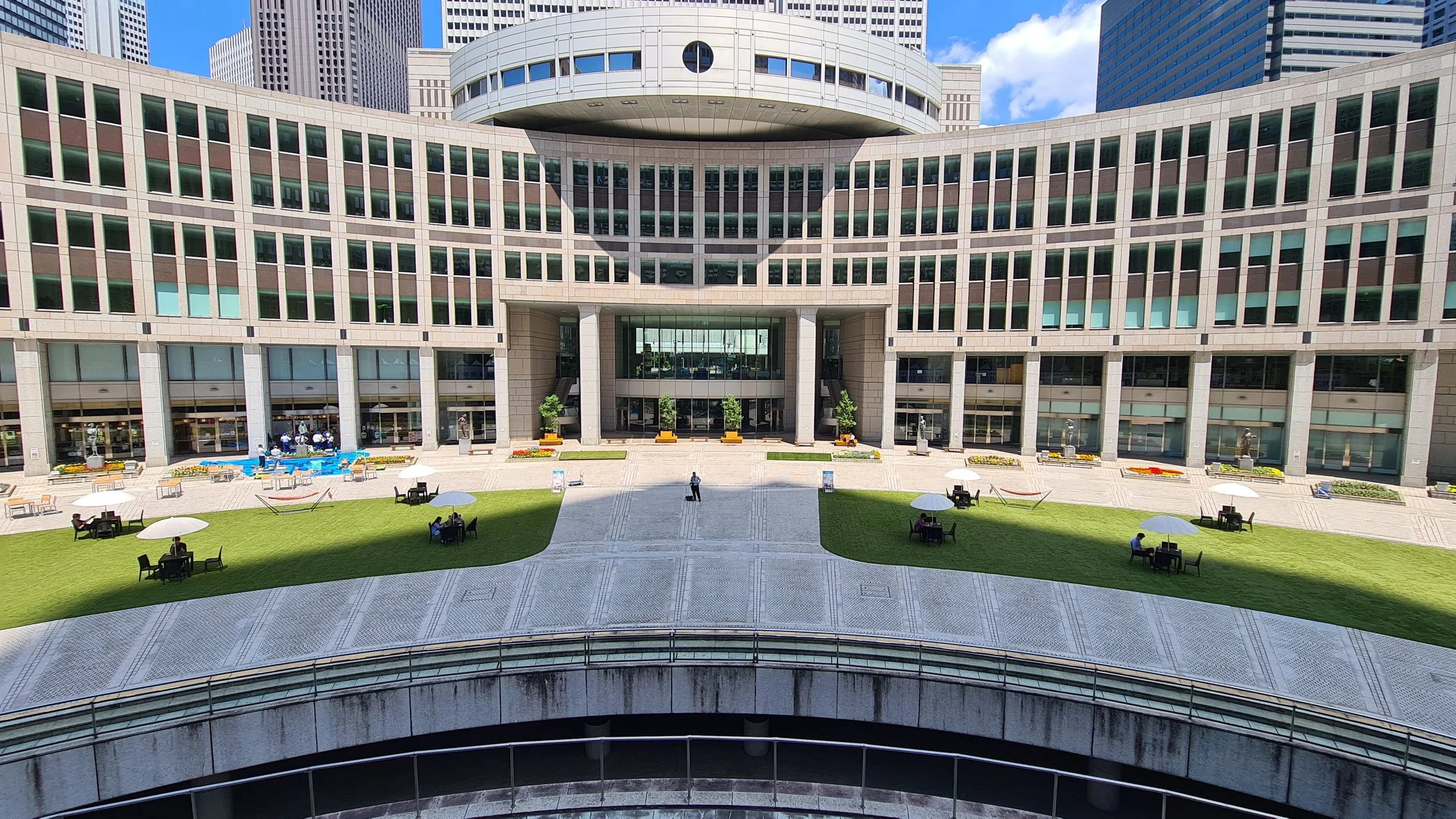 Facade of a modern office building with a plaza area featuring tables with umbrellas, green lawns, and a few people walking or sitting.