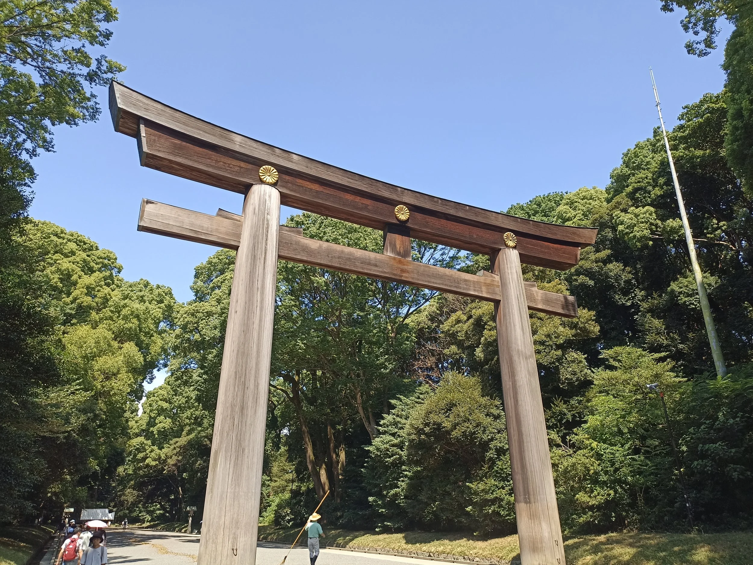 A large wooden torii gate standing outdoors amidst green trees under a clear blue sky, with a few people walking nearby.