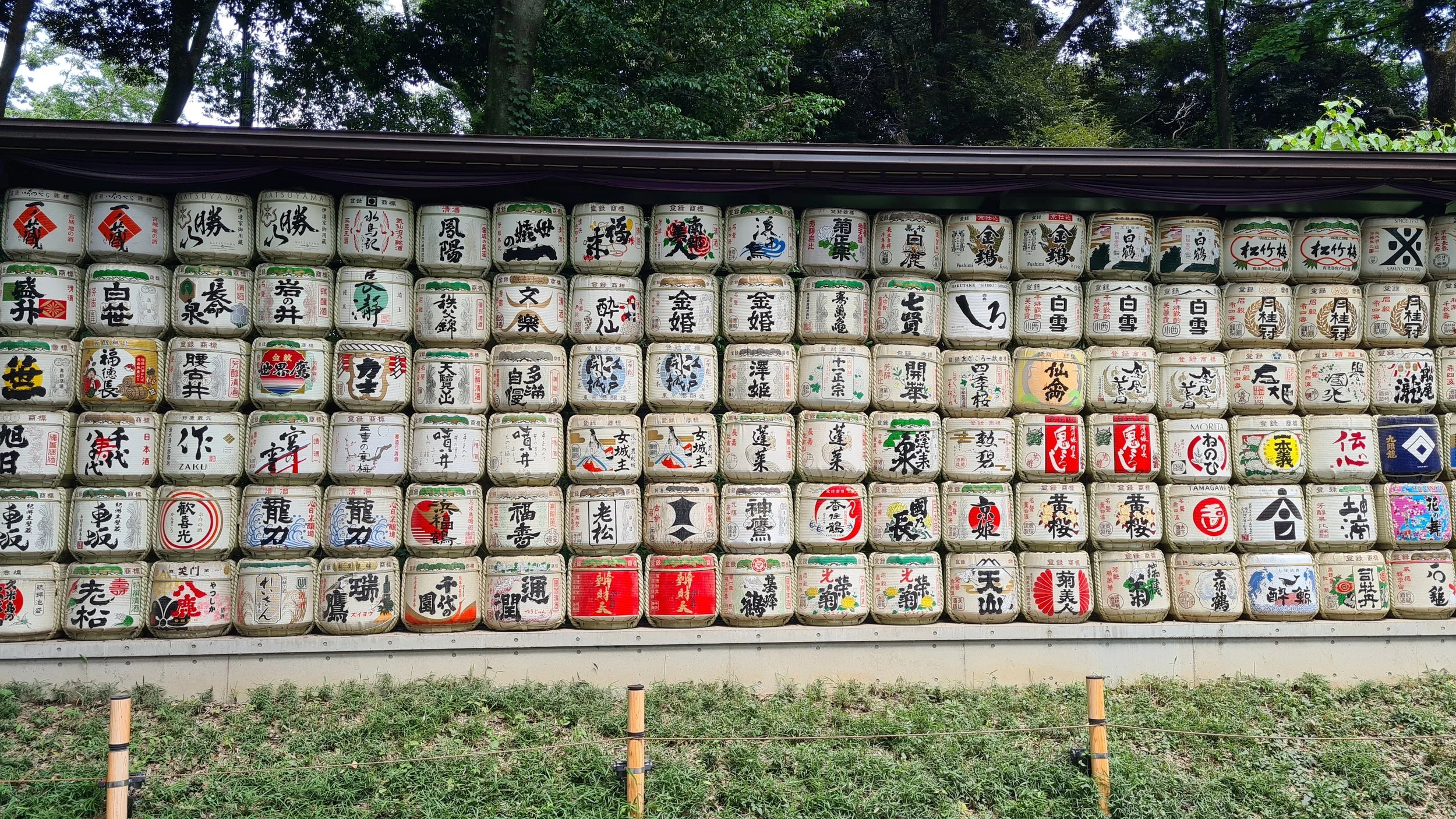A large display of traditional Japanese sake barrels arranged in a grid, with each barrel decorated with various Japanese characters and designs, set outdoors with trees in the background.
