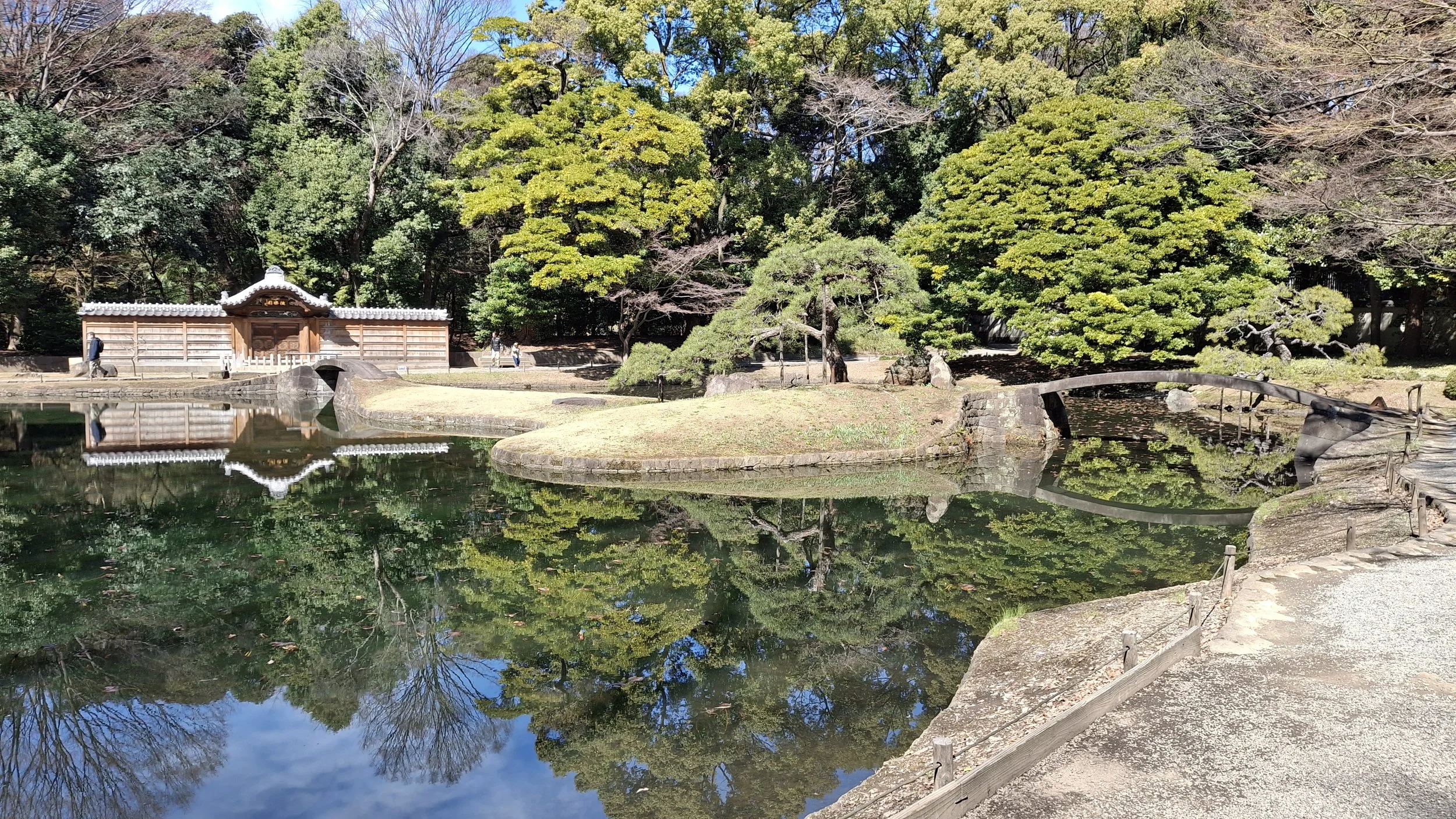 A tranquil Japanese garden with a pond reflecting the trees and sky, a small arched bridge over the water, and a wooden structure with traditional tiled roof in the background, surrounded by lush greenery.