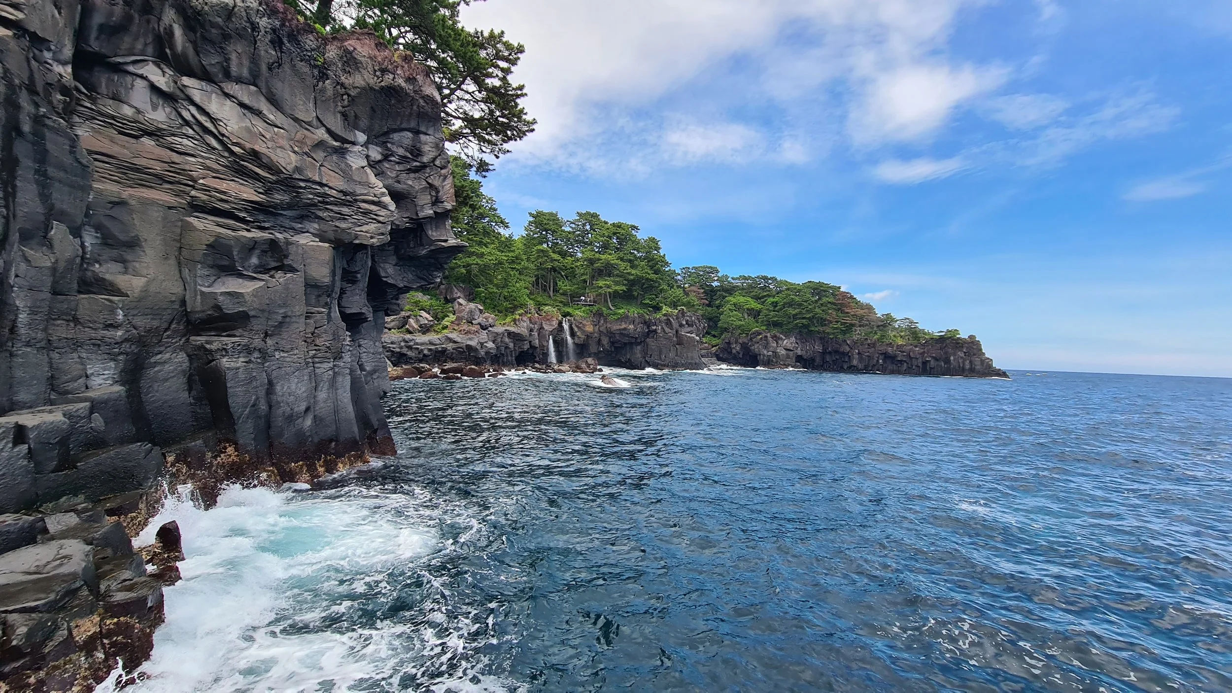 Rocky coastline with cliffs and greenery, blue ocean water, and a partly cloudy sky
