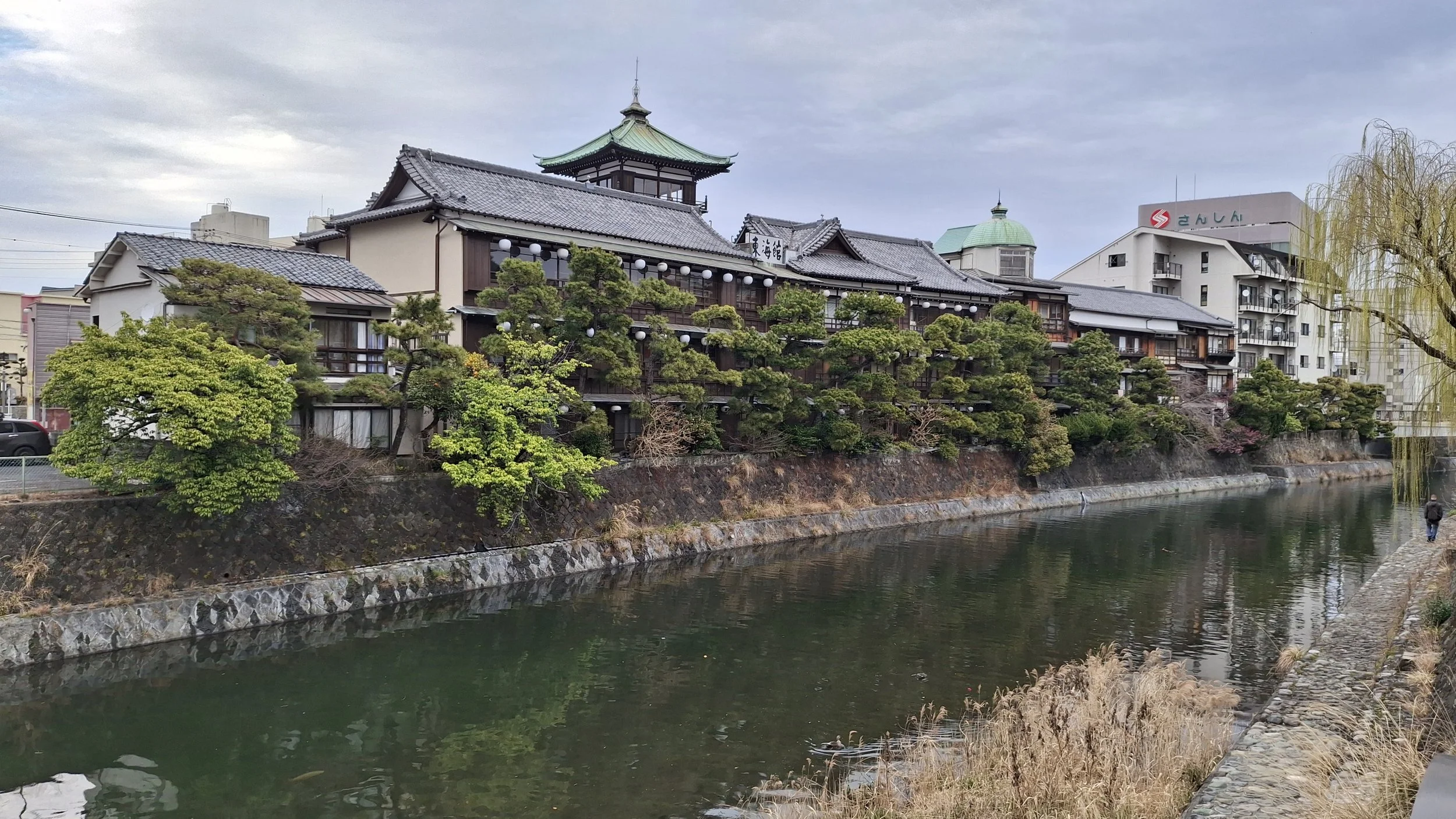Traditional Japanese building with multiple stories, surrounded by lush greenery and trees, next to a calm river with a stone embankment, under a cloudy sky.