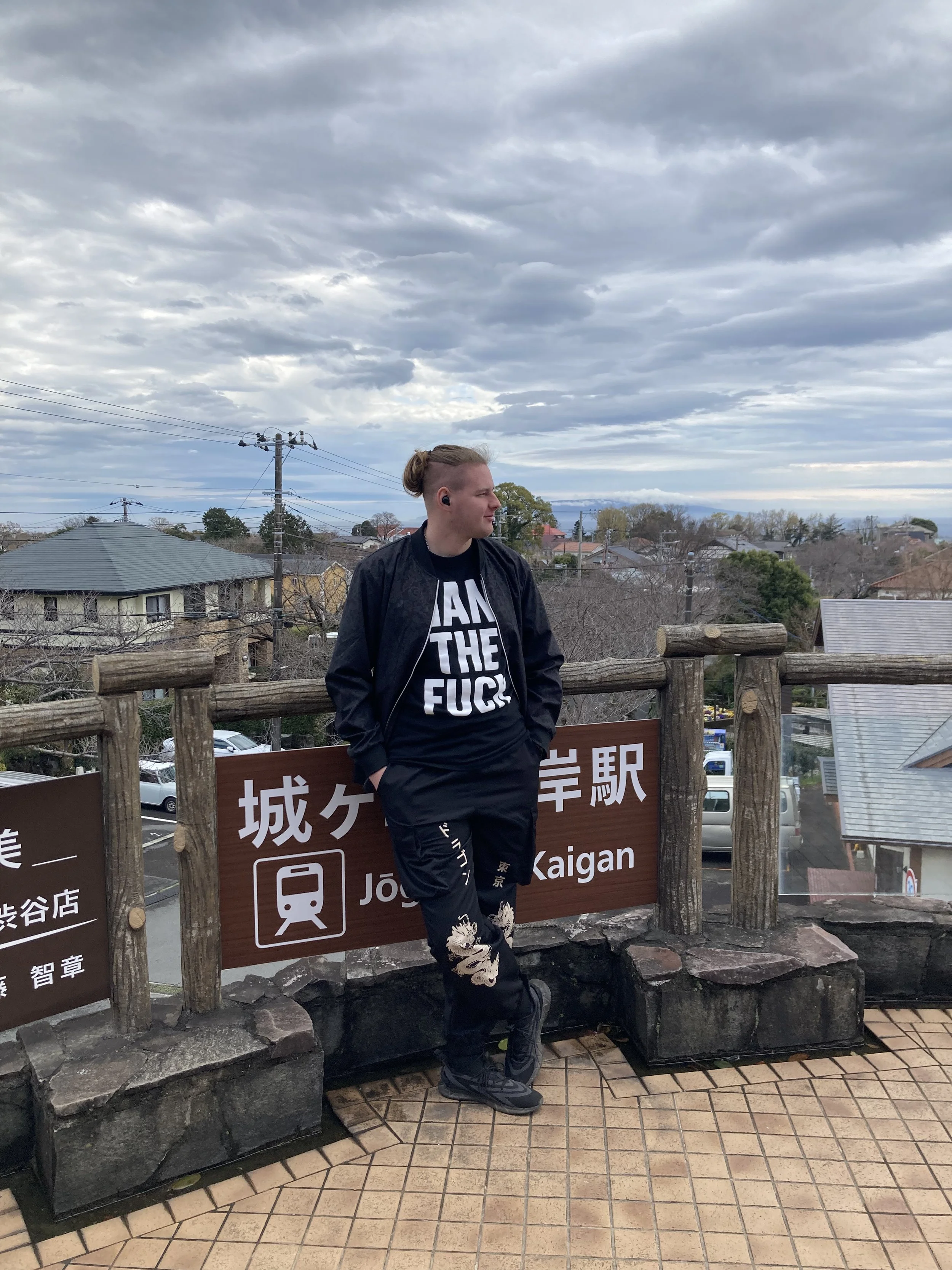 A person with short hair, wearing a black jacket and a black T-shirt with white text, stands on an outdoor observation deck near a sign that says 'Jōban-Kaigan' in both Japanese and English, with a rural town and cloudy sky in the background.