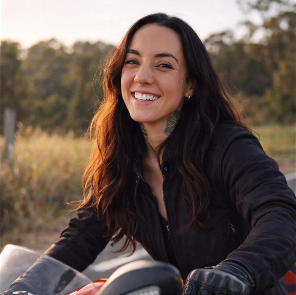 A woman with long dark hair smiling while sitting on a motorcycle outdoors during sunset.