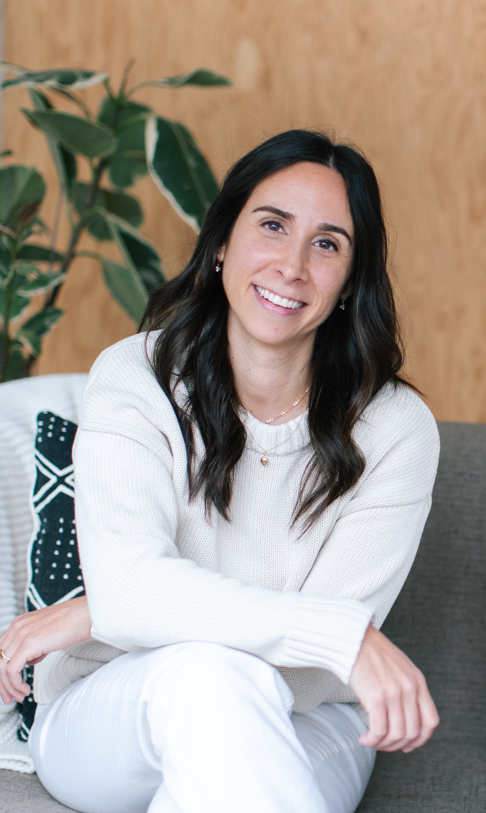 A woman with long dark hair and a friendly smile, wearing a white sweater and jewelry, sitting indoors in front of a wooden wall and a large green plant.