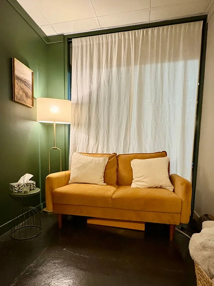 Yellow loveseat with two white pillows, next to a floor lamp and a small side table with a tissue box, in front of cream curtains.