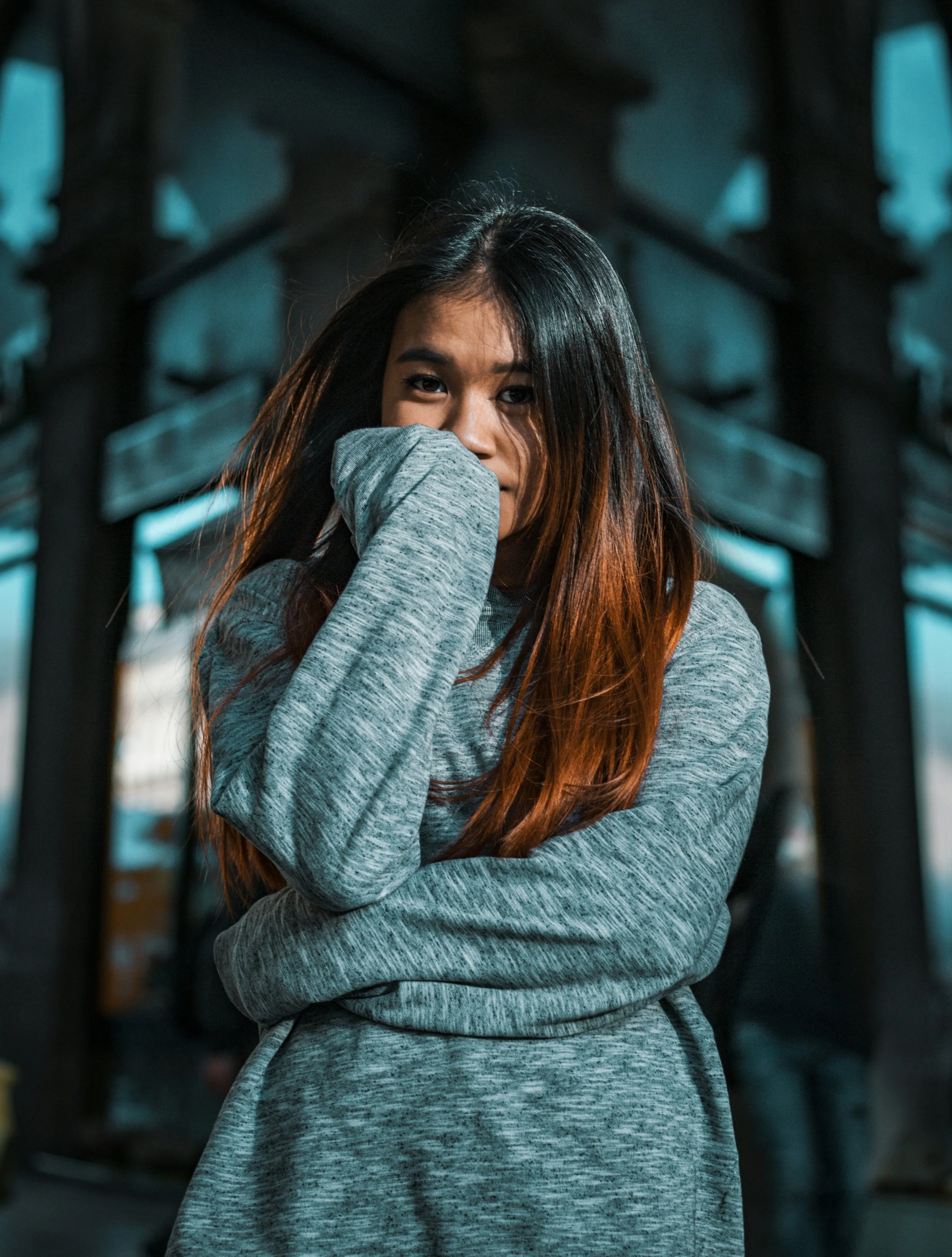 A young woman with long dark hair and tan highlights, wearing a gray long sleeve shirt, covering her mouth with her hand while standing outdoors near a black iron fence during daytime with moody lighting.