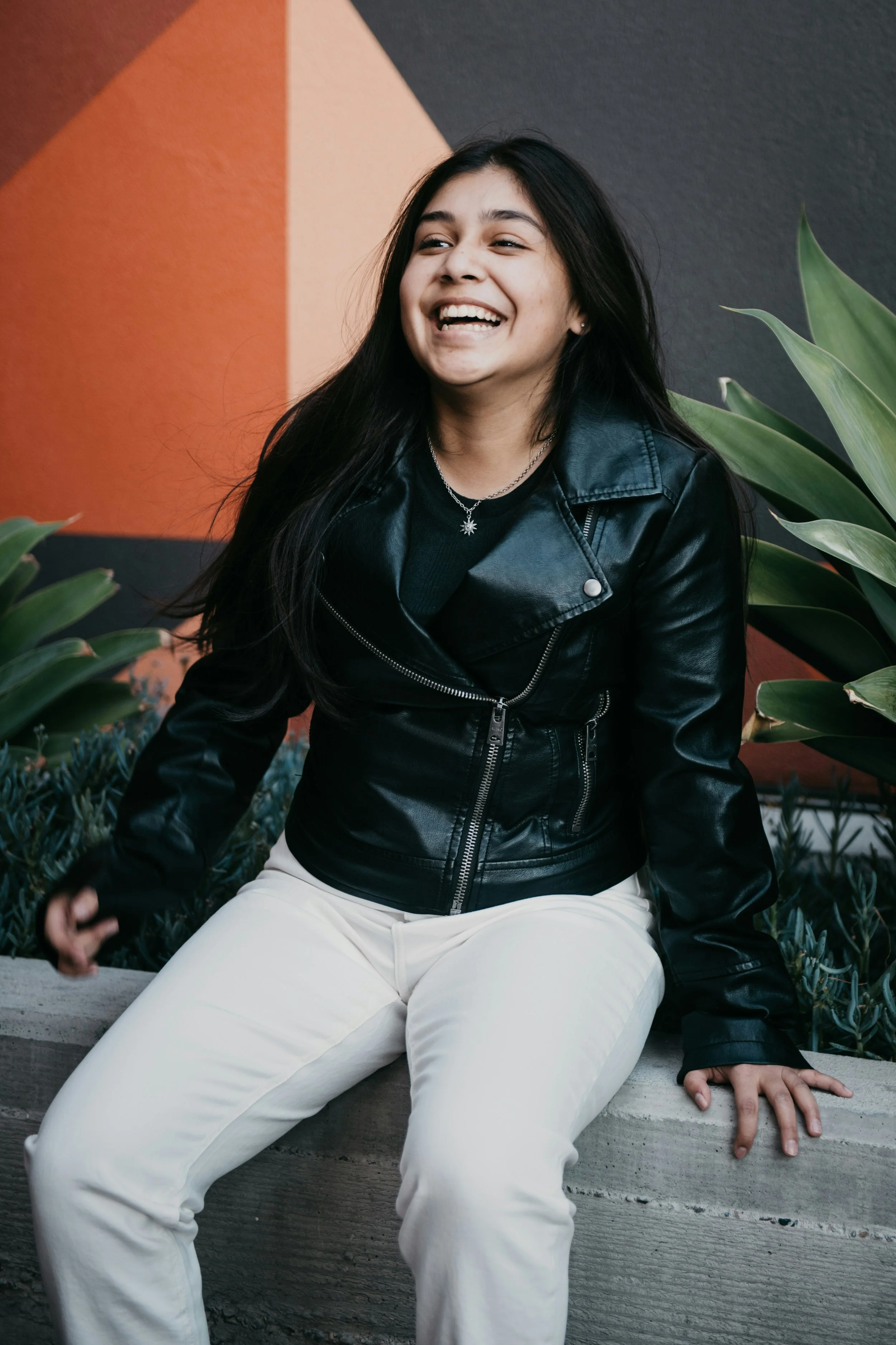 A young woman with long black hair, wearing a black leather jacket and white pants, sitting on a concrete ledge in front of a colorful geometric wall and lush green plants, smiling and looking happy.