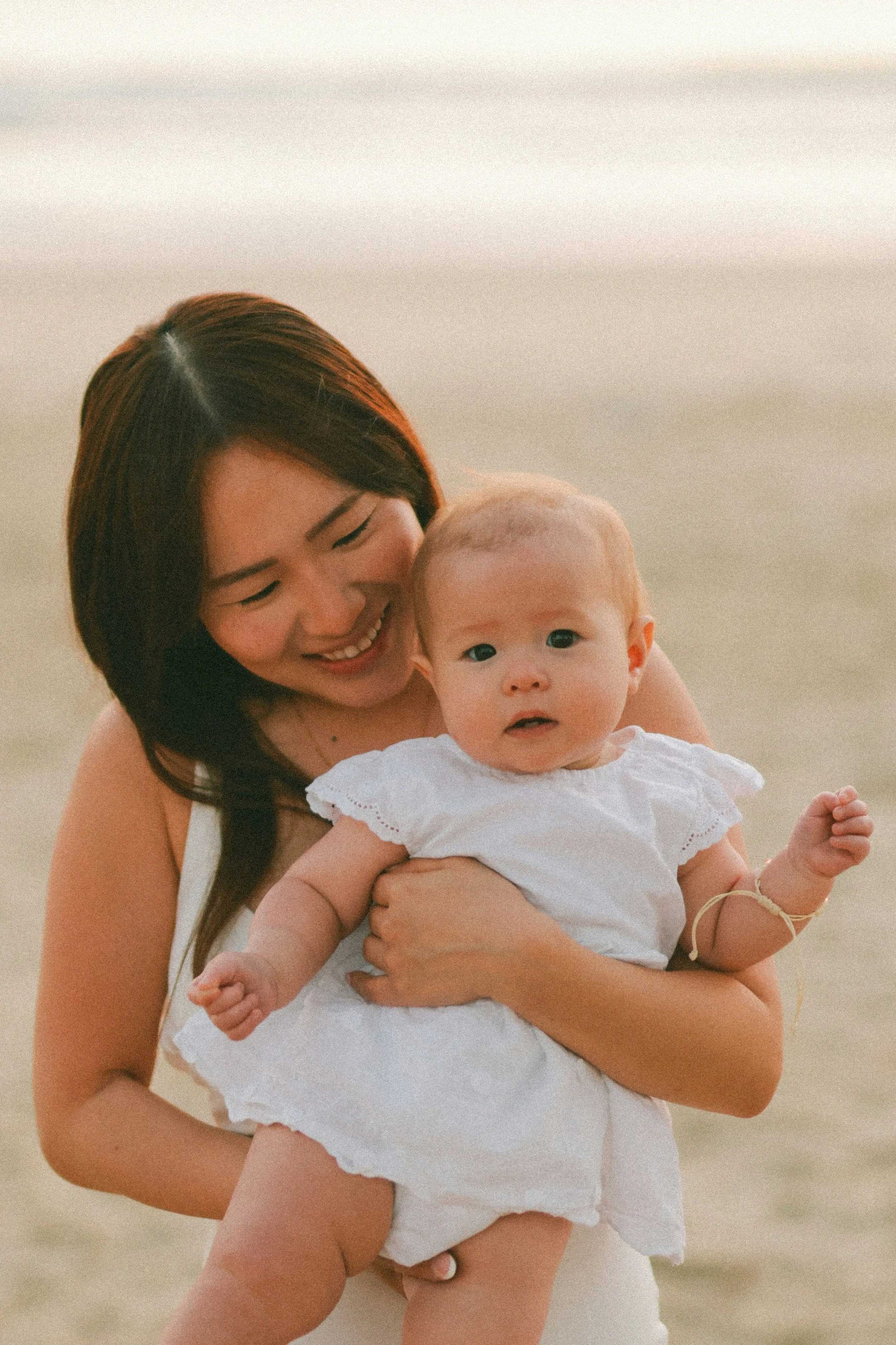A woman holding a baby at the beach during sunset, both smiling and looking happy.