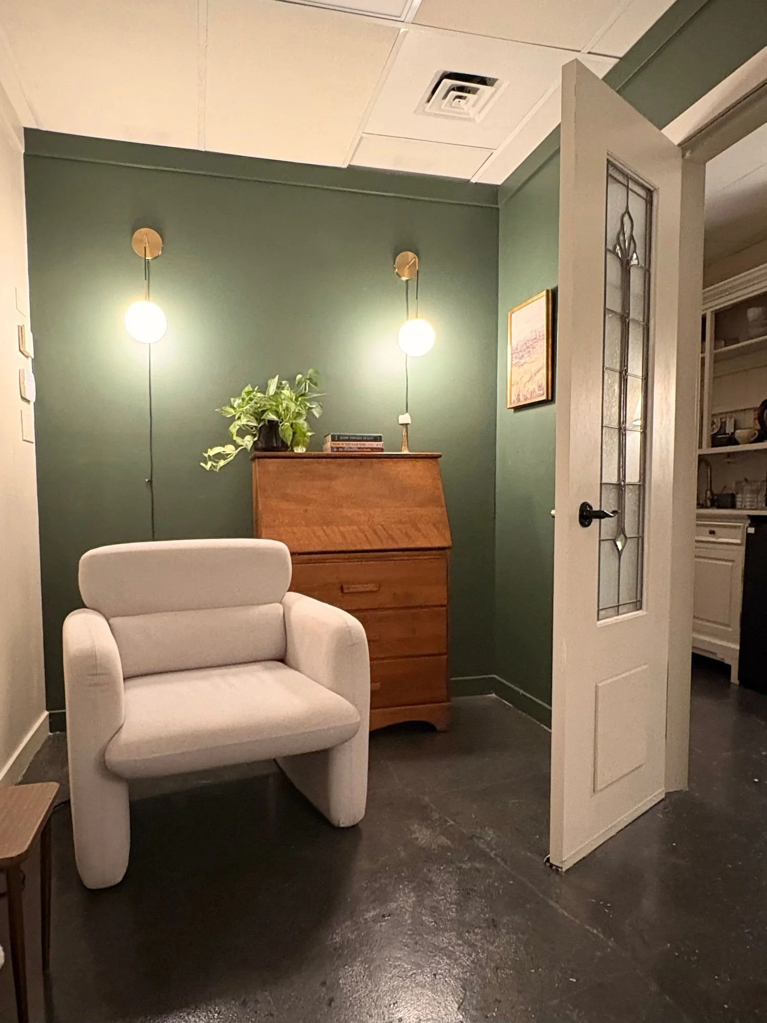 A cozy living room corner with a white armchair and a green wall. Above the dresser with books and a potted plant, two pendant lights hang. An open door with decorative glass panels leads to another room.