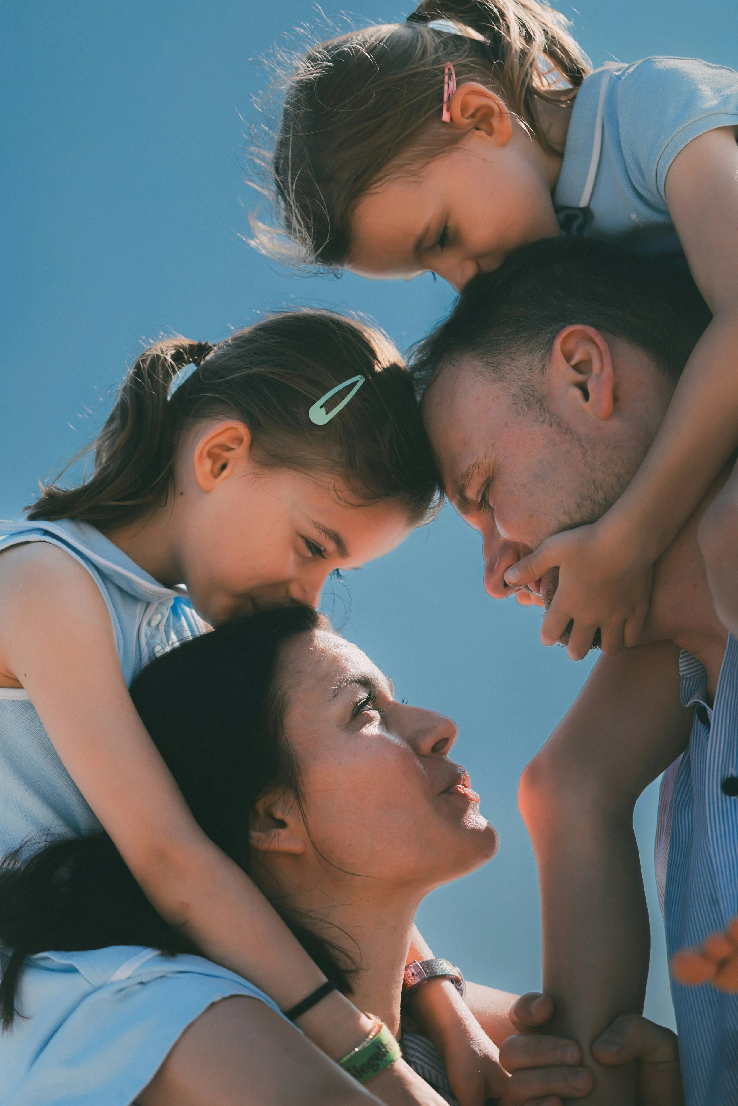 A group of children and a woman are engaged in an outdoor activity. The woman is holding two children upside down, and they are all smiling and looking at each other. The sky is clear and blue.