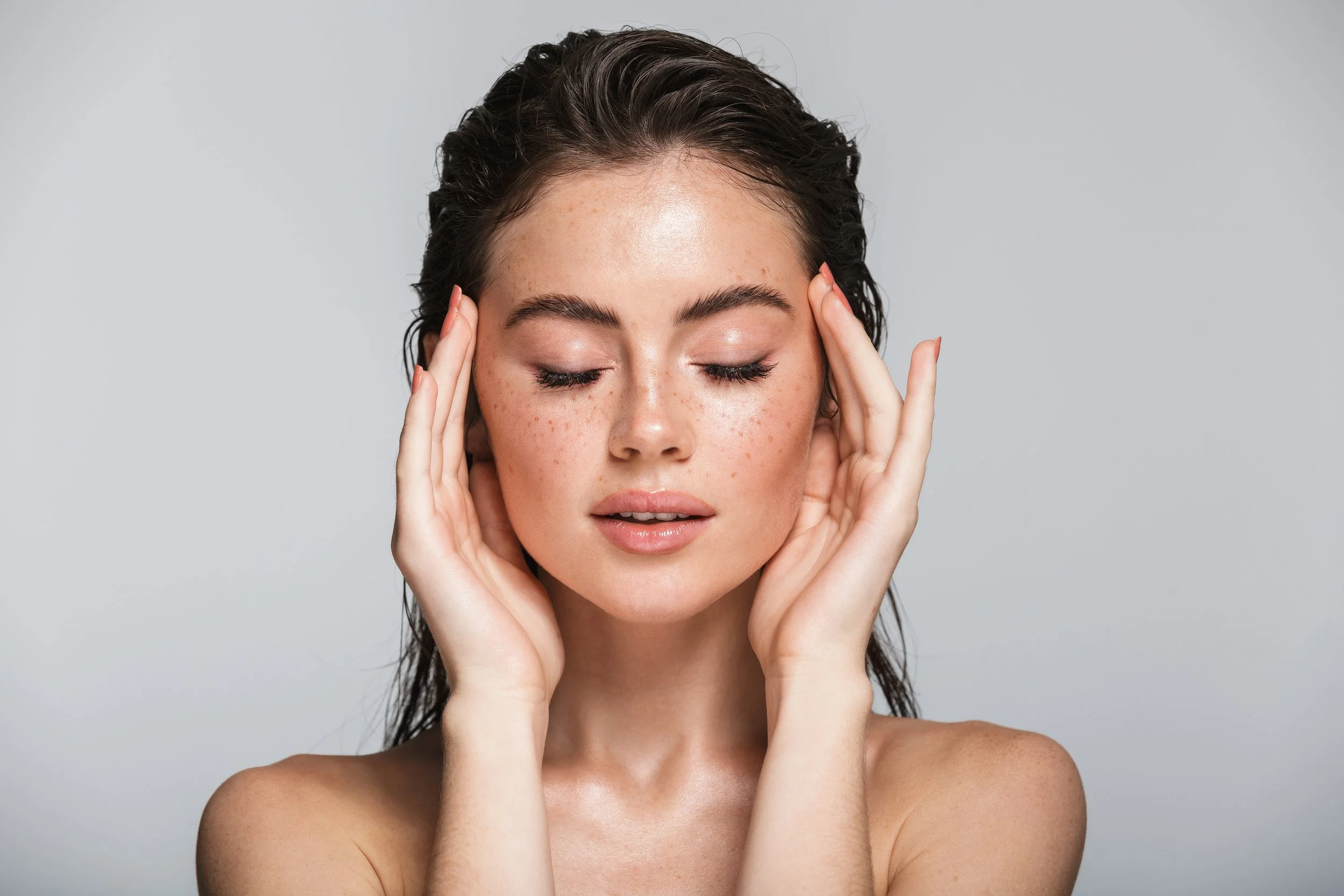 A young woman with damp hair, freckles, and natural makeup, with her hands touching her temples and eyes closed against a plain gray background.