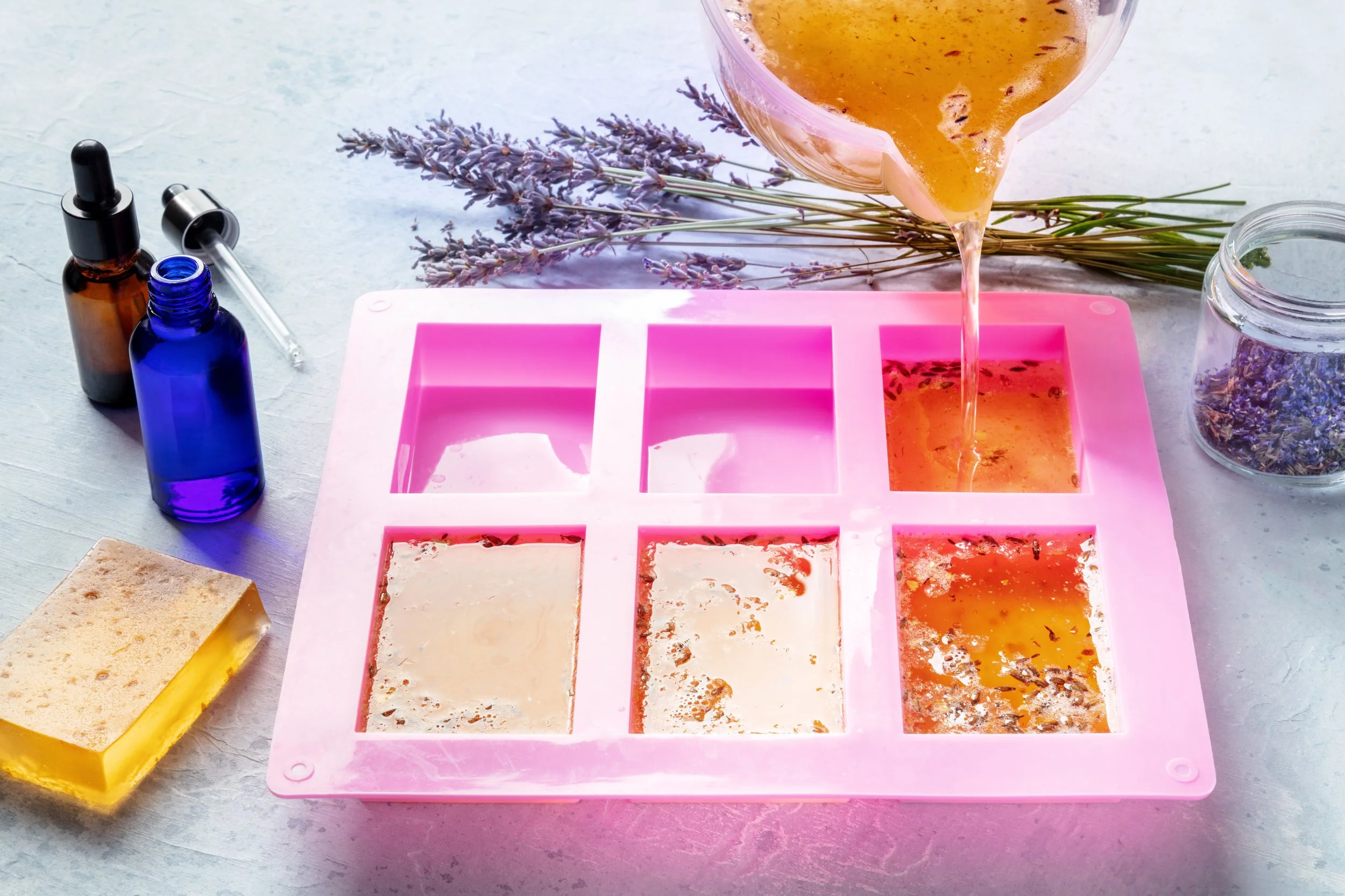 Pouring lavender-infused liquid into an ice cube tray with lavender sprigs and soap on a textured surface.