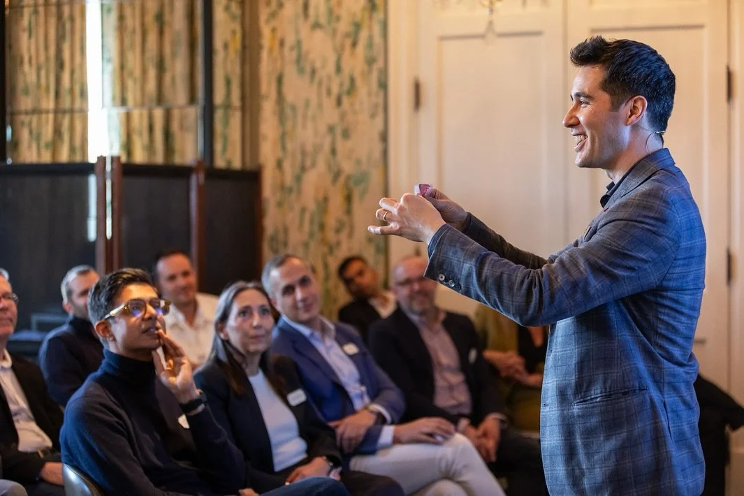 David Kwong in a gray plaid suit giving a presentation to an audience in a decorated room.