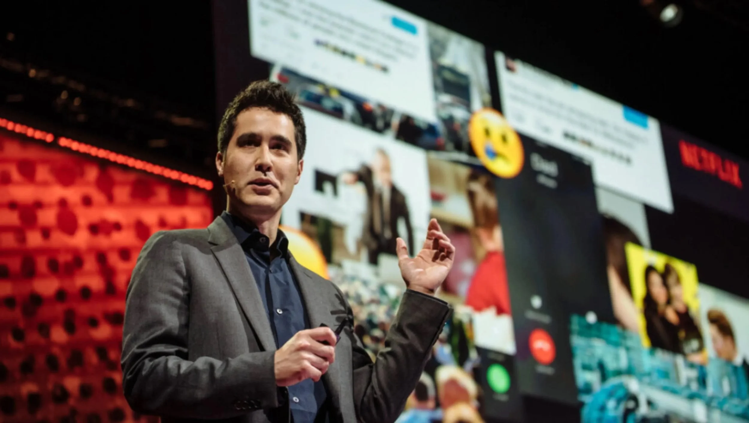 David Kwong in a gray suit and blue shirt speaking on stage at TED with a microphone, gesturing with his right hand, behind giant screens displaying social media posts and a yellow emoji with a winking face with tongue out.