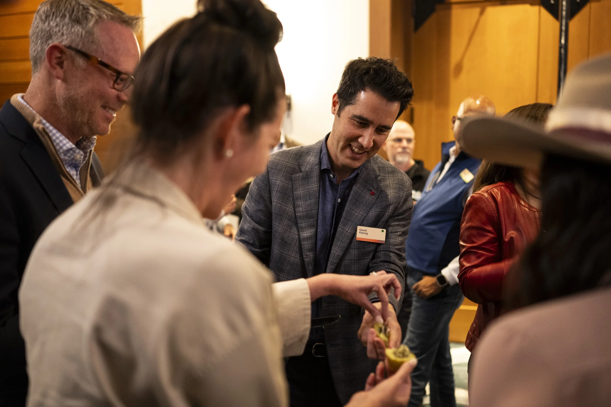 People at a social gathering, with David Kwong in a grey checkered blazer smiling and helping others with food.