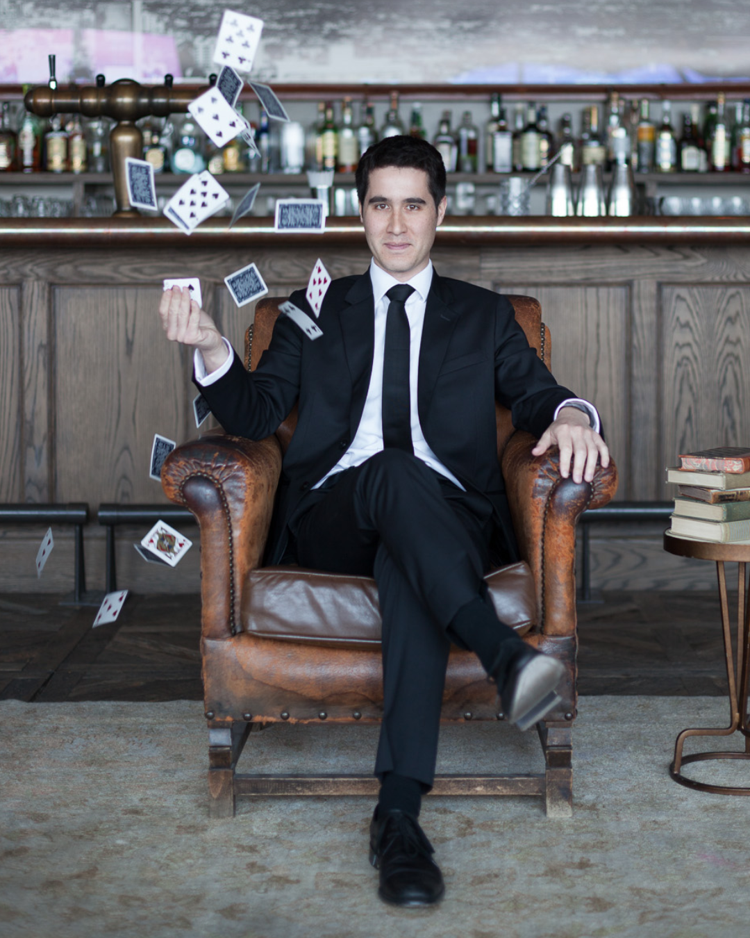 David Kwong in a black suit and tie sitting in an armchair with playing cards flying around him in a bar setting.