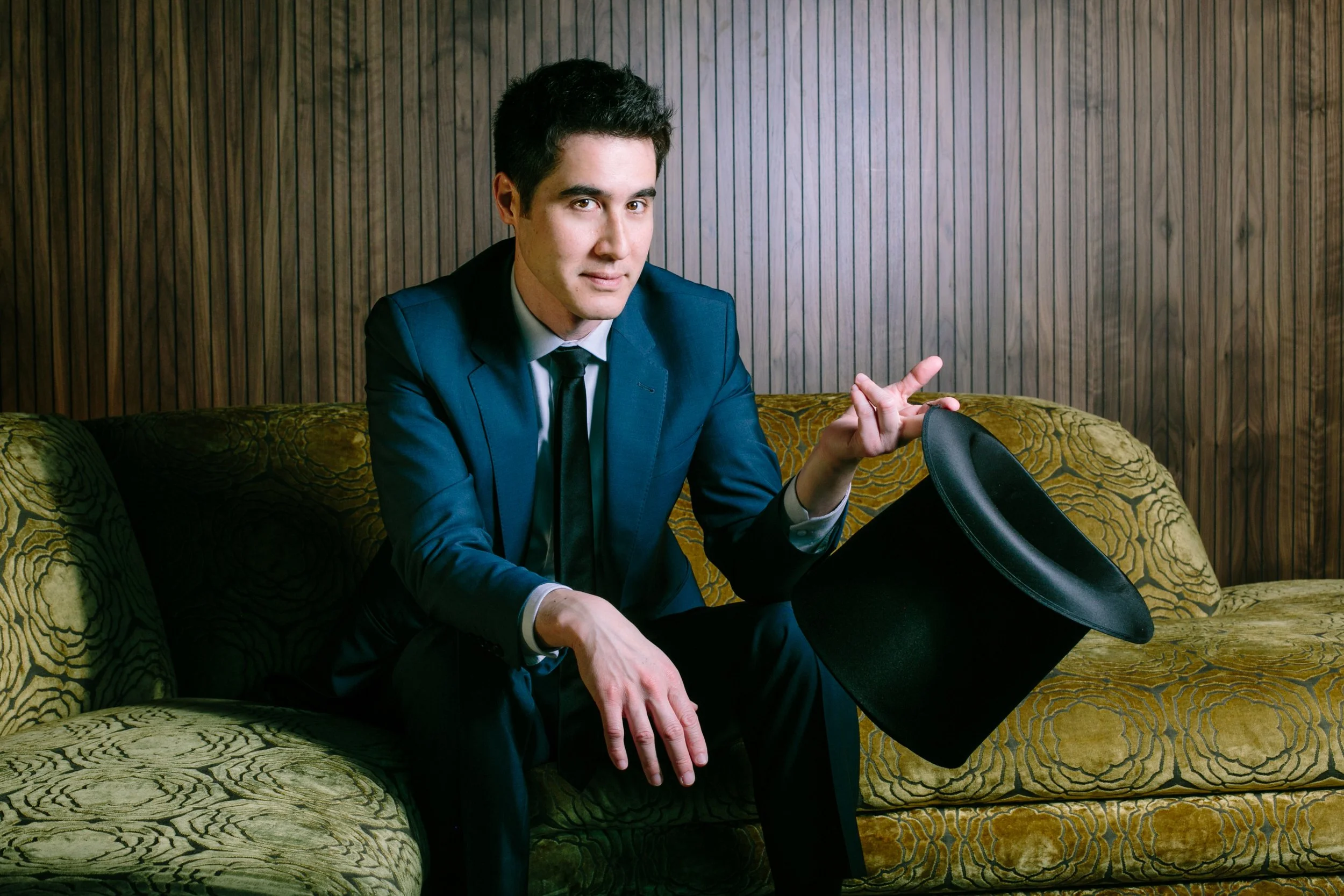 David Kwong in a blue suit sitting on a vintage green patterned couch holding a black top hat, with a wooden-paneled wall background.