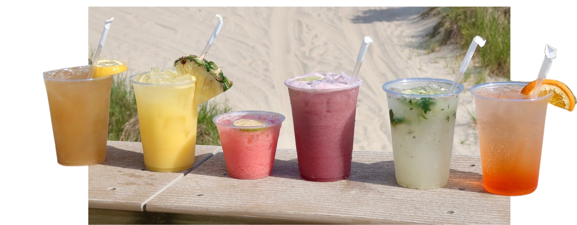 Six colorful tropical drinks with straws lined up on a wooden surface, with a sandy beach and grass in the background.