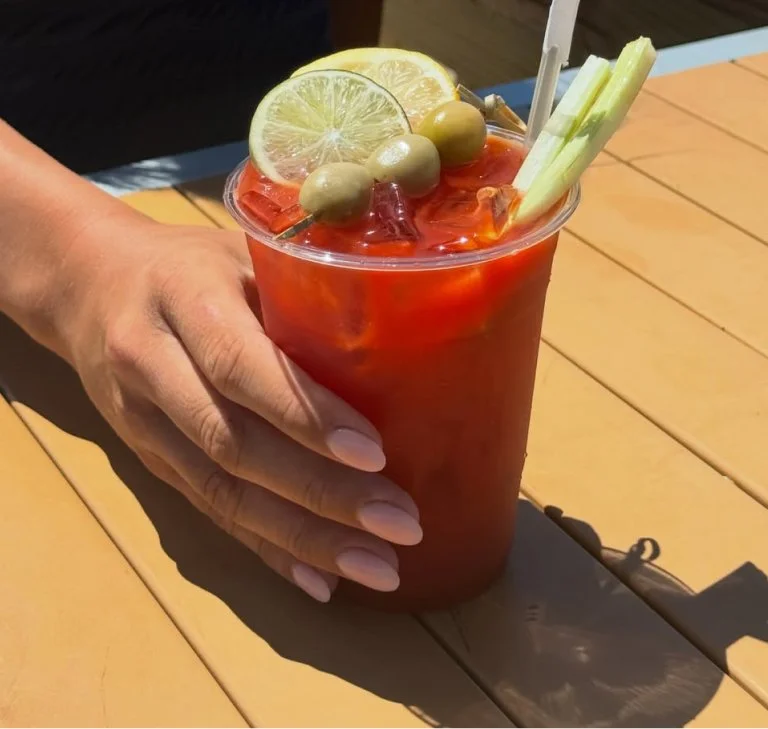 A hand holding a tall glass of bloody Mary garnished with a lemon slice, green olives, and a celery stick