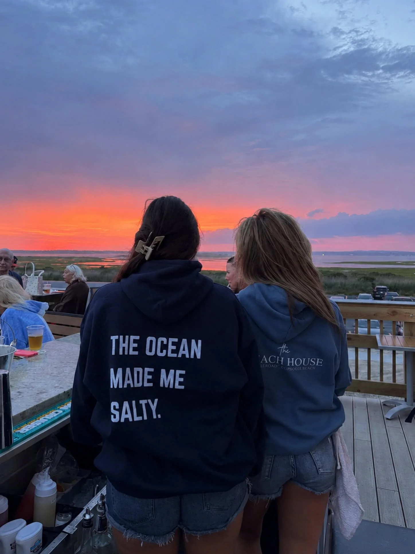People gathered on a deck at sunset, with a view of a river or bay and colorful sky.