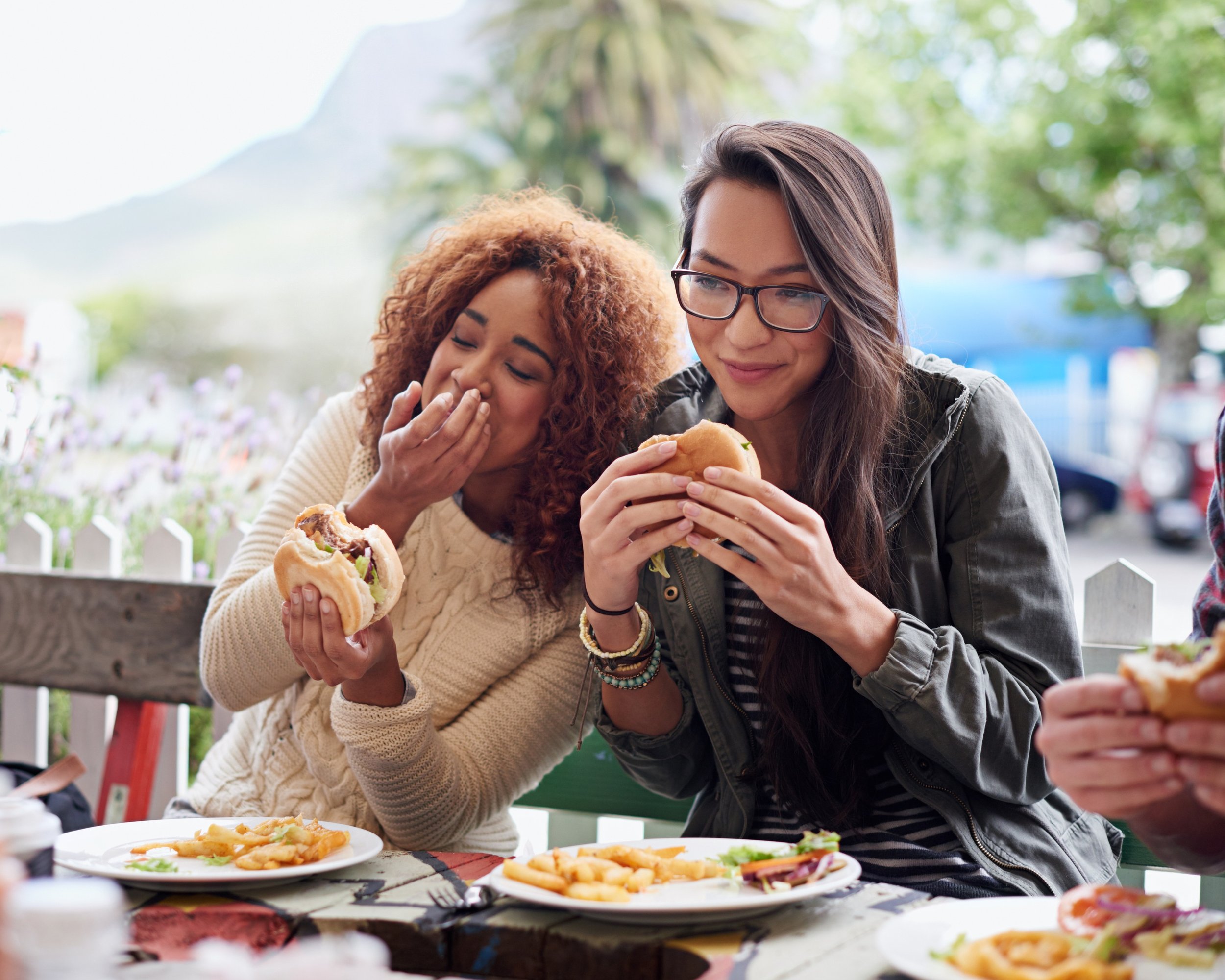 Two women enjoying food outdoors. One woman with curly hair and a cream sweater is eating a burger, and another woman with straight hair and glasses is holding a burger. Plates of fries and other food are on the table.