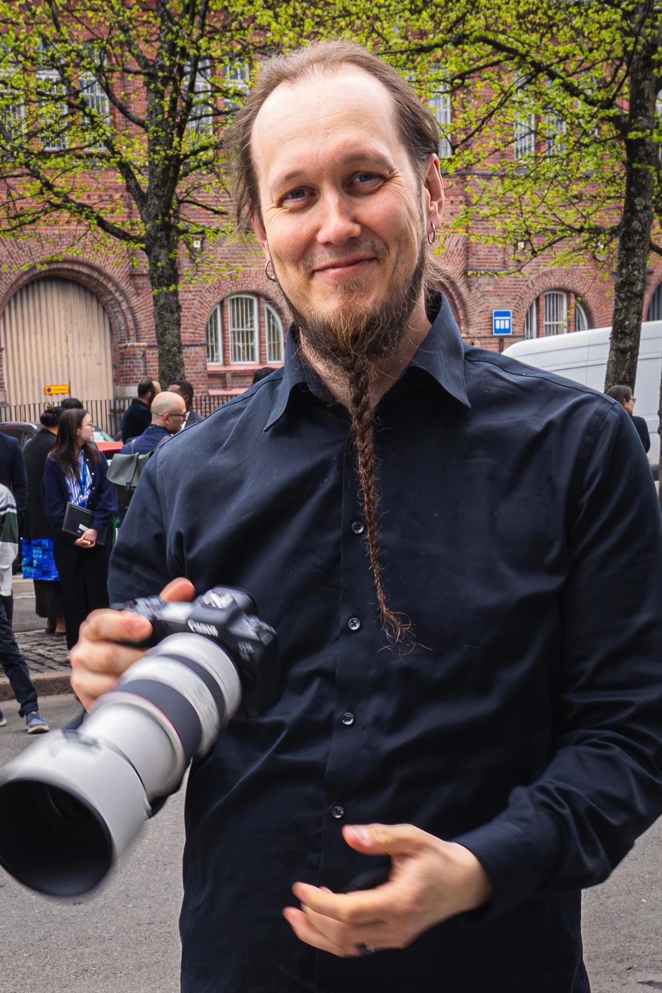 Mies ihailee kameraa ja pitää sitä kädessään valokuvia ottaessaan, taustalla on ihmisiä ja rakennuksia puistossa tai kadulla.