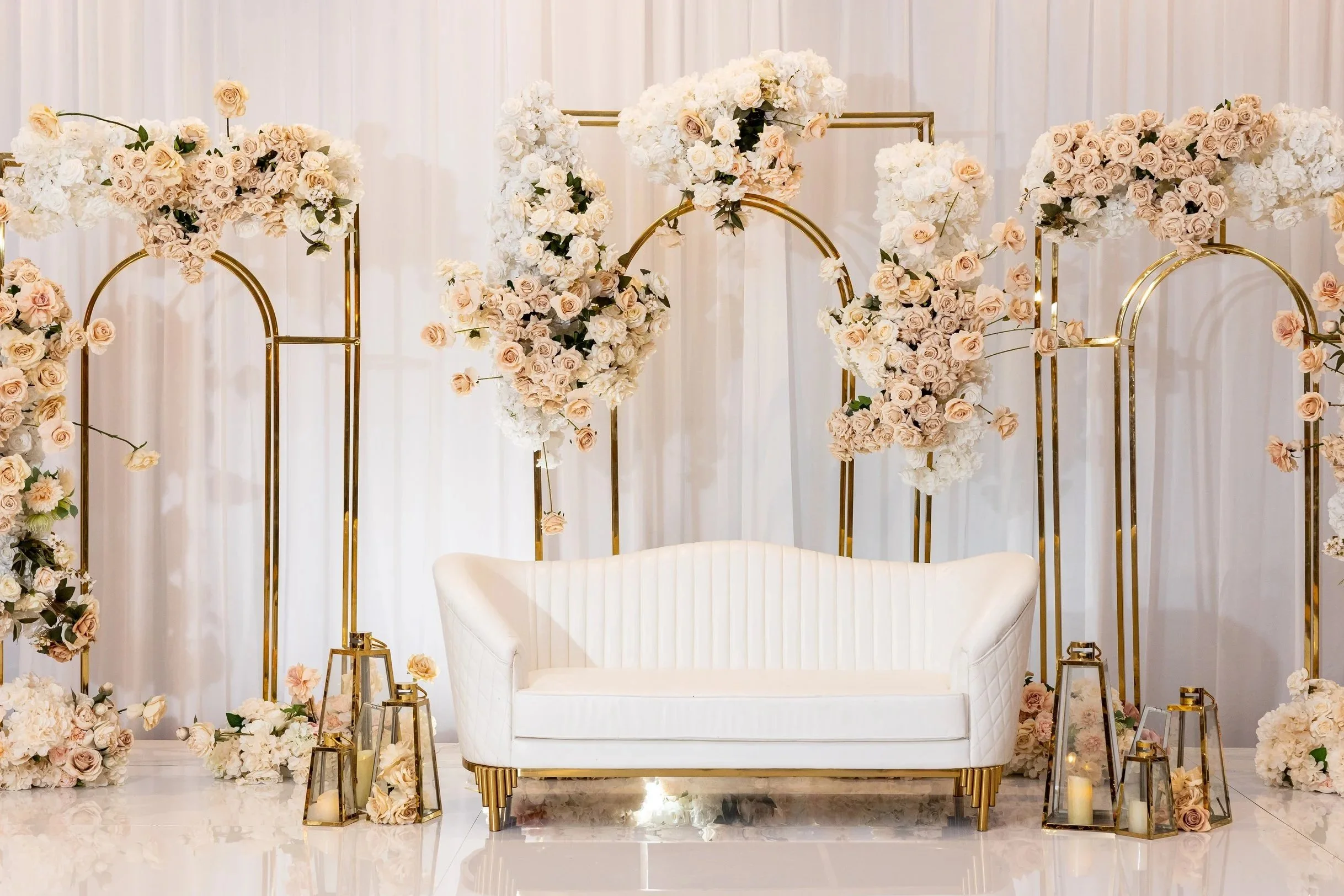 Elegant wedding backdrop decorated with white and blush pink roses and hydrangeas, gold geometric frames, and lanterns with candles, with a white curved sofa in front.