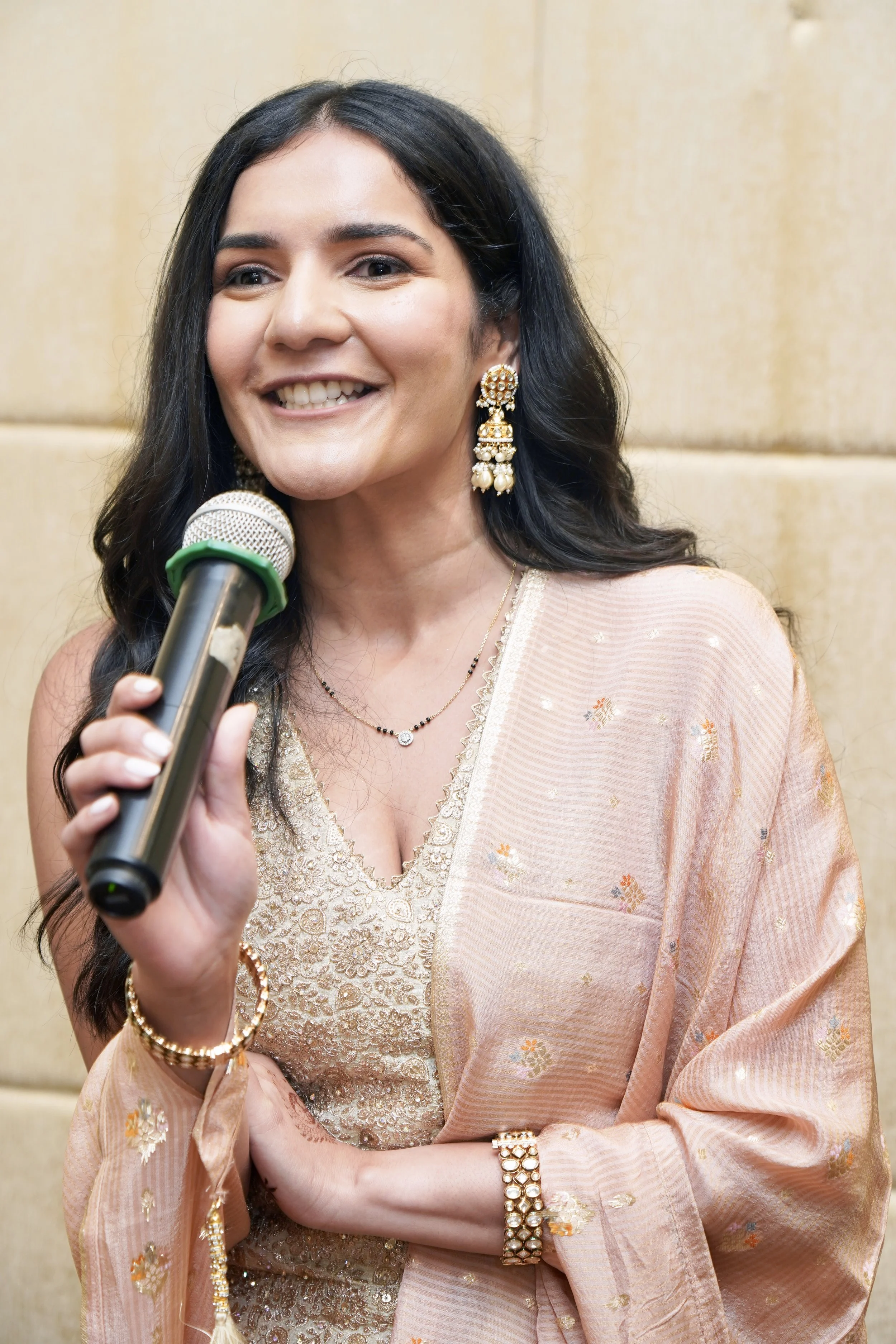 A woman in traditional Indian attire, holding a microphone, smiling, wearing large earrings, a necklace, a bracelet, and a pink embroidered saree, standing against a beige wall.