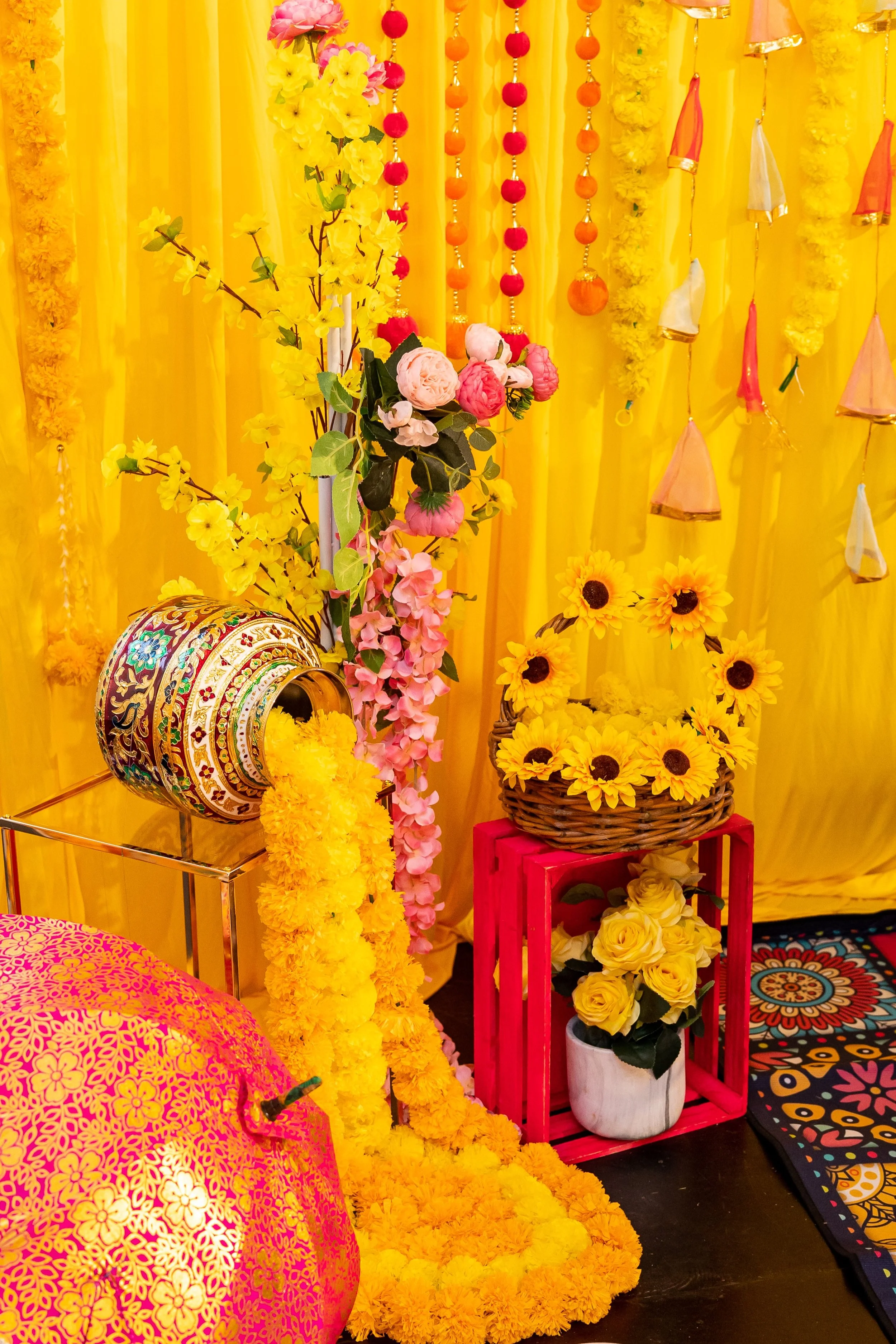 Decorative setup with yellow and pink flowers, yellow marigold garlands, sunflower and yellow roses, colorful hanging ornaments, and vibrant textiles.