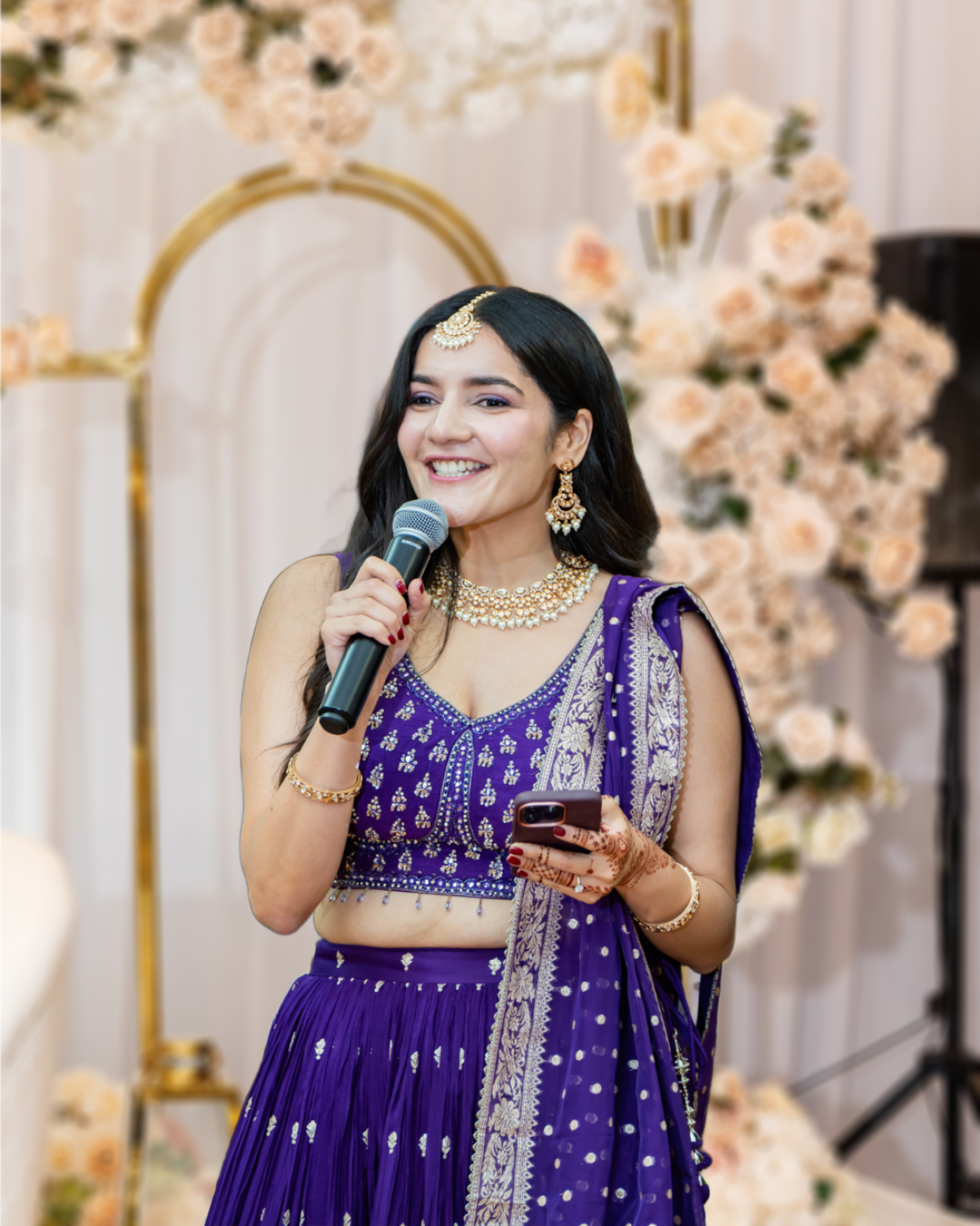 A woman dressed in traditional Indian attire, holding a microphone and a phone, at a decorated event with flowers in the background.