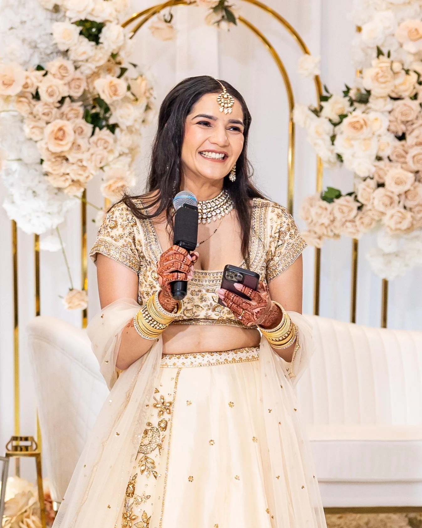 A woman in traditional Indian attire smiling and holding a microphone and phone, surrounded by floral decorations.