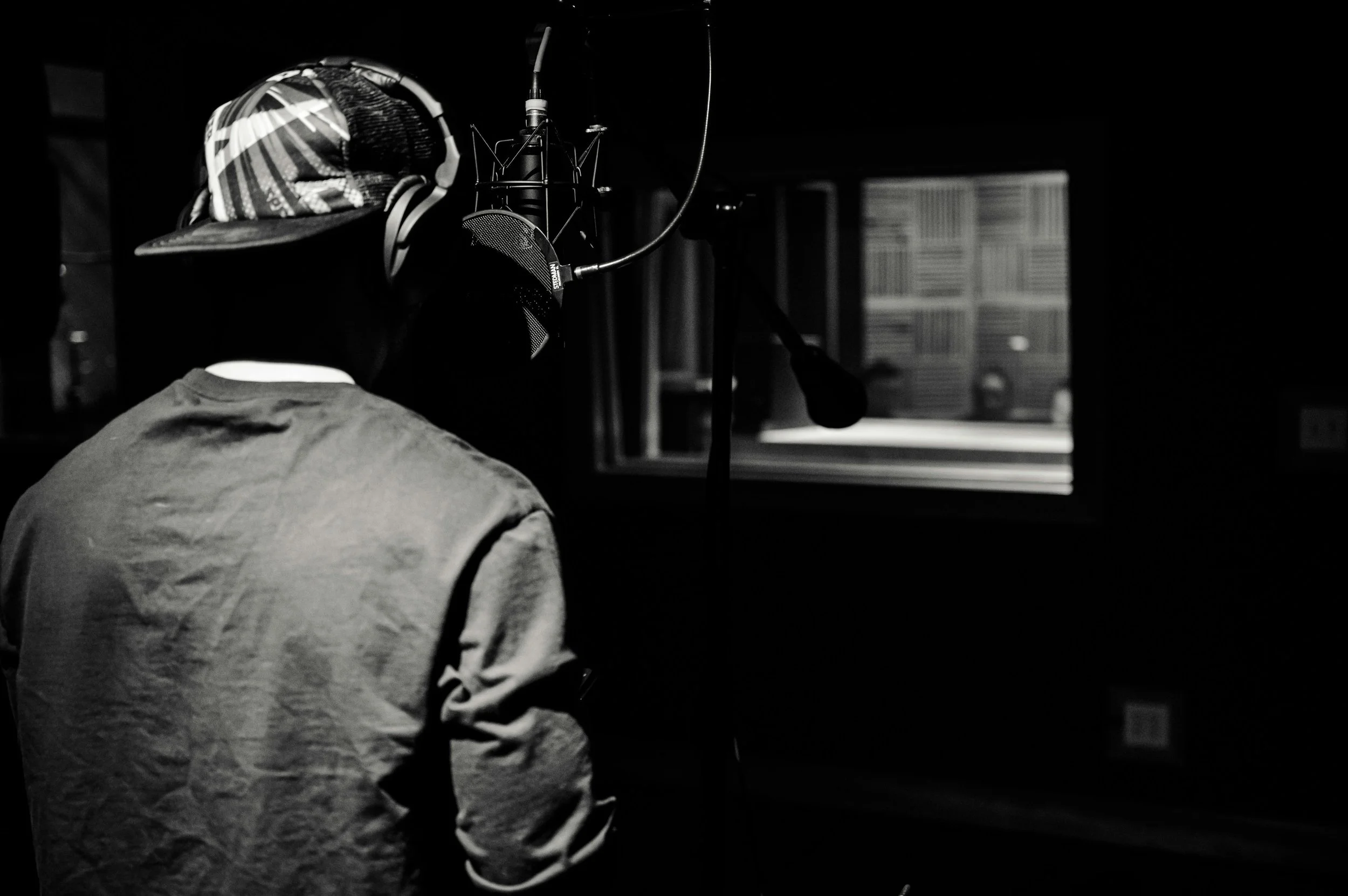 A person wearing a cap and headphones is in a recording studio, singing or speaking into a microphone. The person is facing a window, looking into an isolation booth or control room.