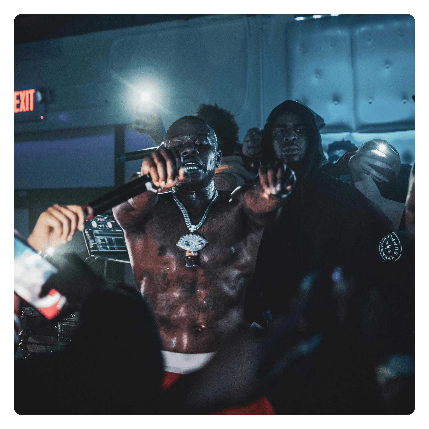 A shirtless man wearing a large chain necklace and holding a microphone, performing passionately in a dimly lit indoor venue with a blue hue, with another person standing nearby.