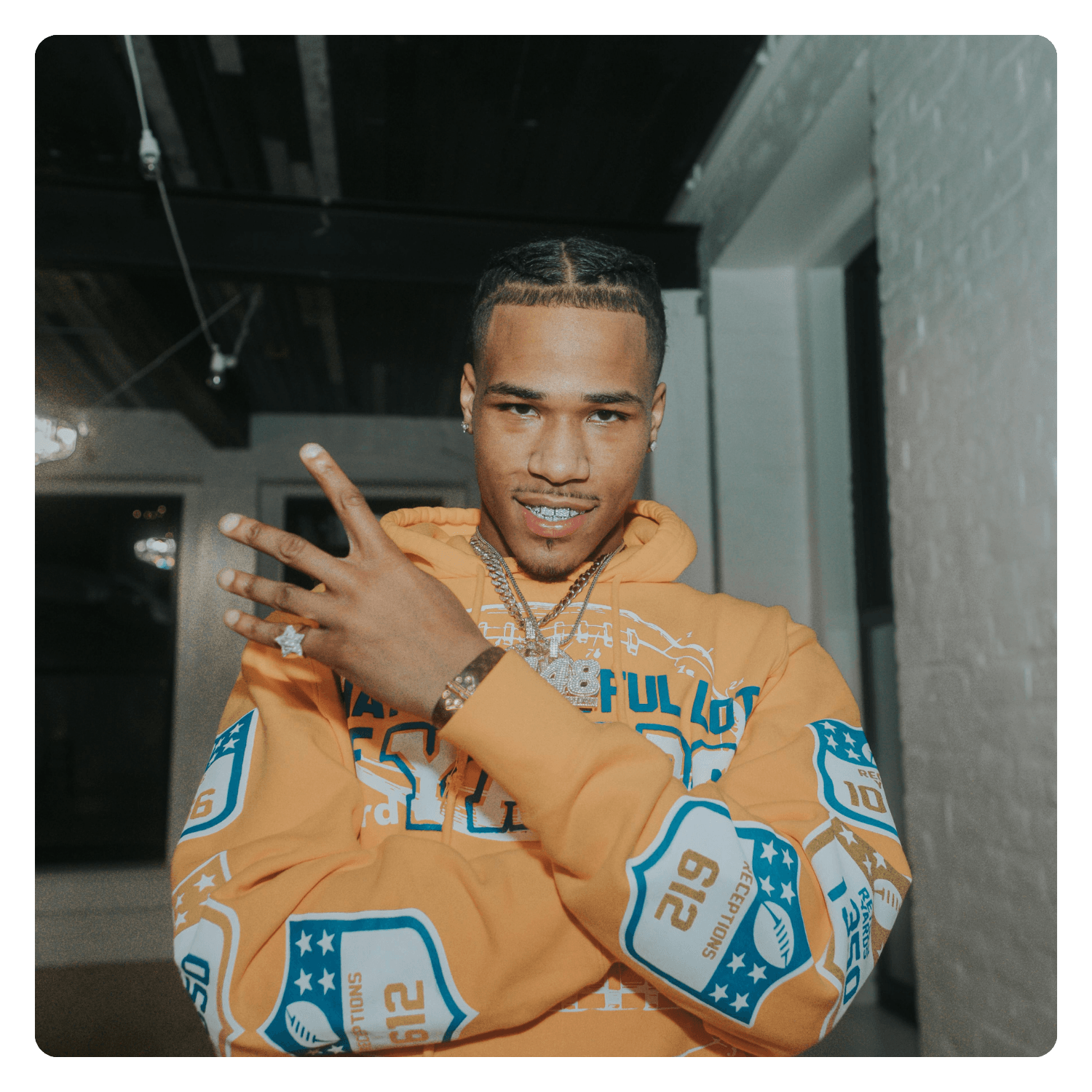 Young man with braided hair wearing a bright orange hoodie and jewelry, posing indoors against a white brick wall.