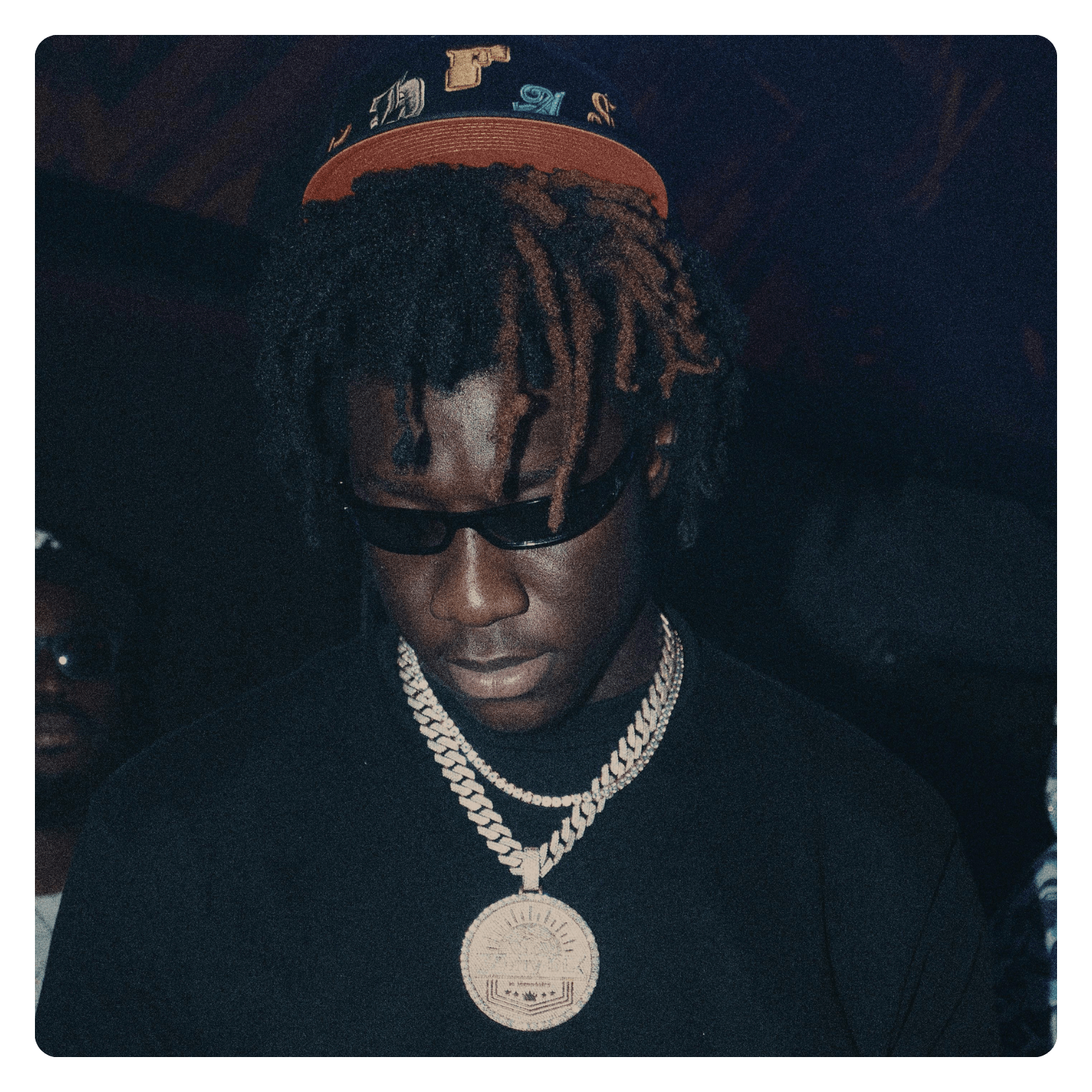 A young man with dreadlocks wearing sunglasses, gold chains, and a black shirt, looking downward with a neutral expression.