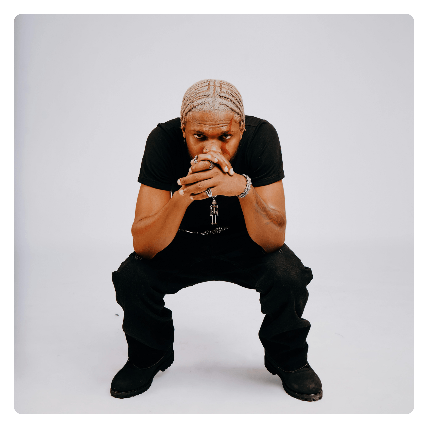 A young man with blonde braided hair, wearing a black t-shirt, black pants, and black boots, squatting with his hands clasped in front of his face against a plain white background.
