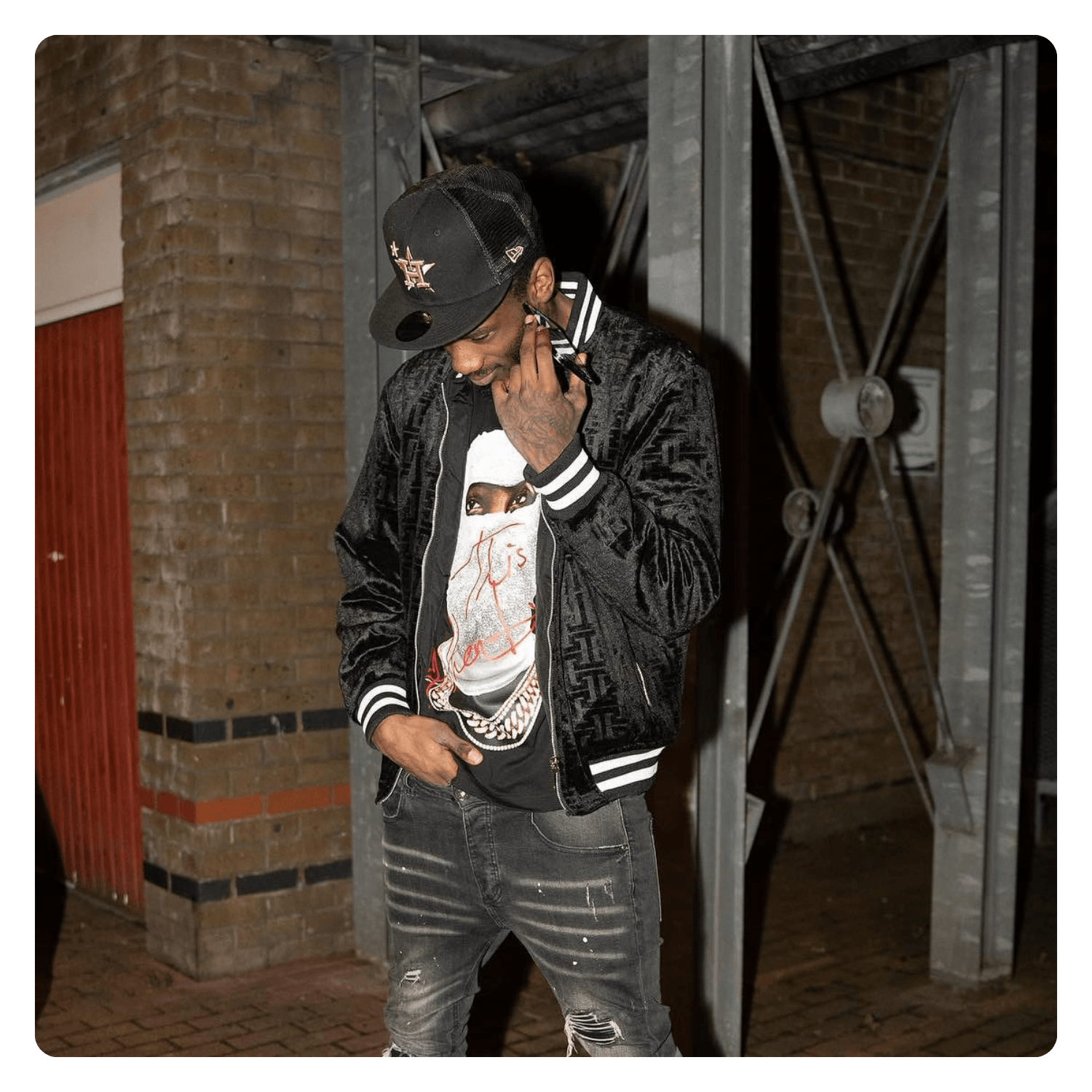 A young man wearing a black baseball cap, black bomber jacket with white striped cuffs, distressed black jeans, and a graphic t-shirt with a woman's face, stands outdoors near a brick wall and metallic structure, smiling and looking down.