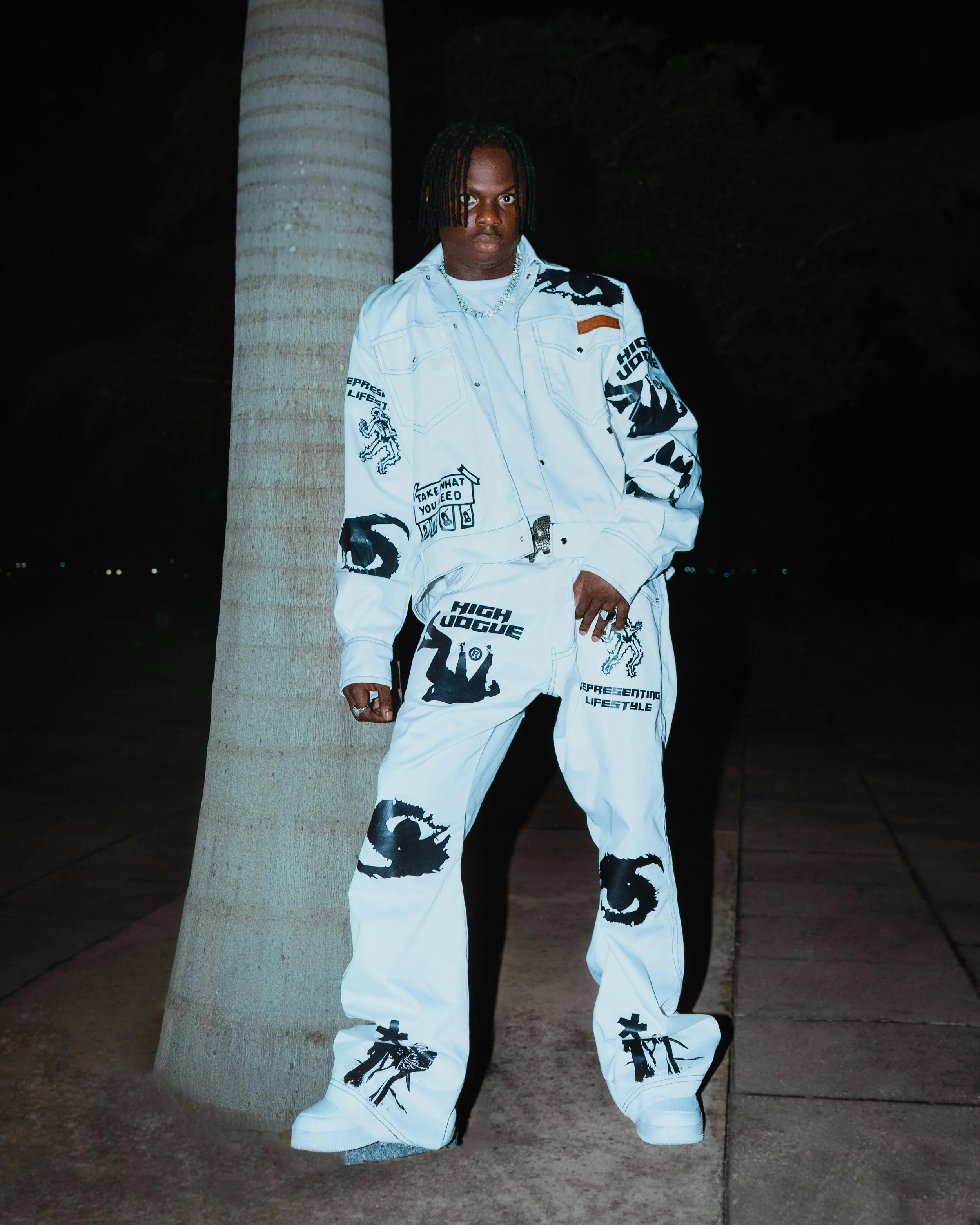 A person standing outdoors at night next to a palm tree, wearing a white graphic jacket and pants with black prints, and white sneakers.