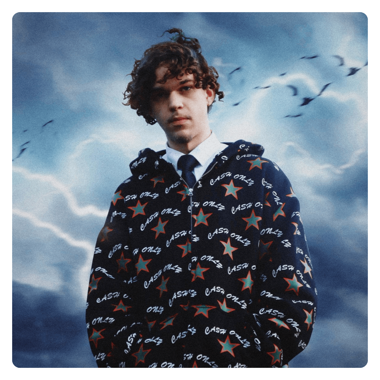 Young man with curly hair standing outdoors against a cloudy sky and a flock of birds flying in the background, wearing a hoodie with a star pattern and the words "CASH ONLY" printed on it, over a collared shirt and tie.