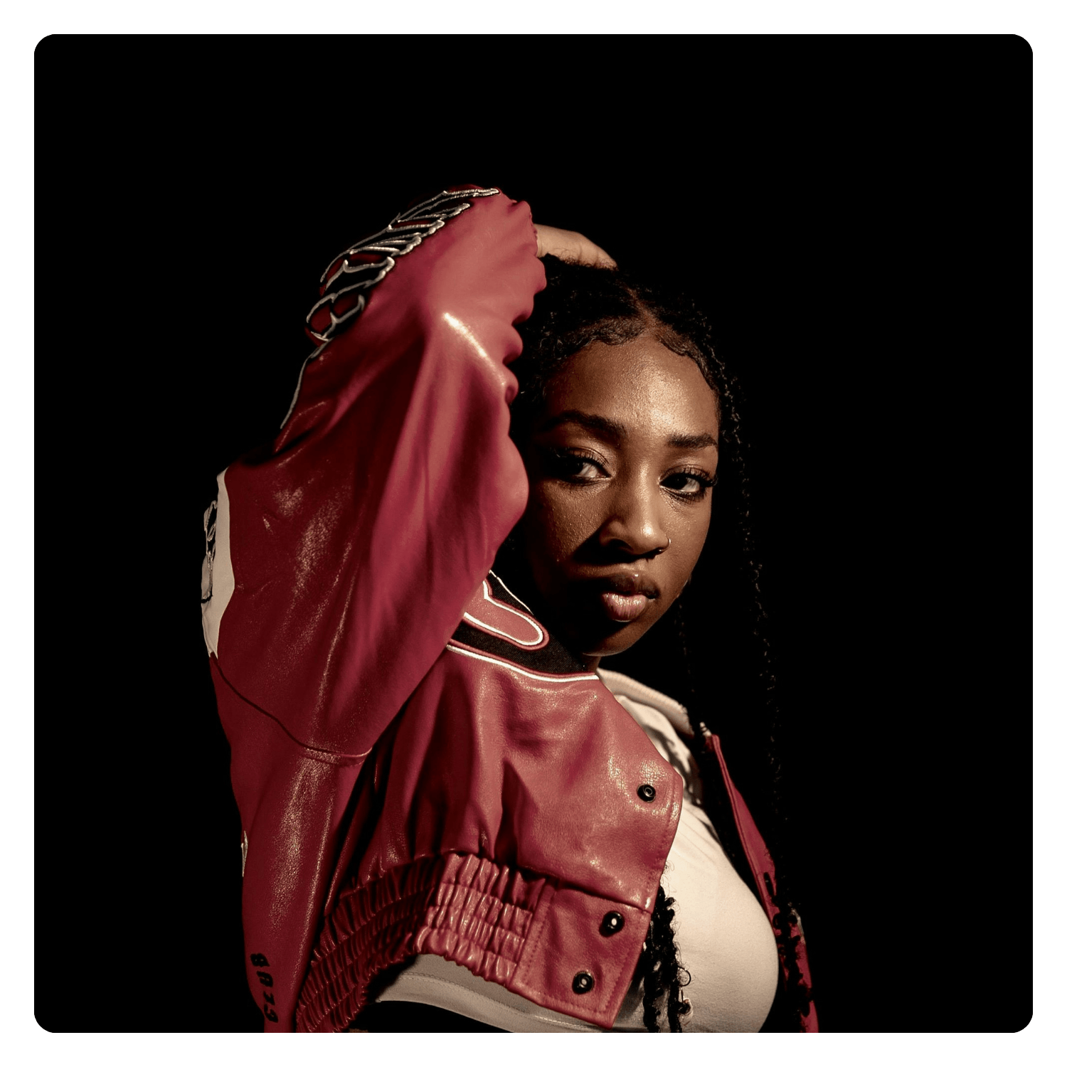 A young woman with dark curly hair poses with one arm raised, wearing a red leather jacket and a white top, against a black background.